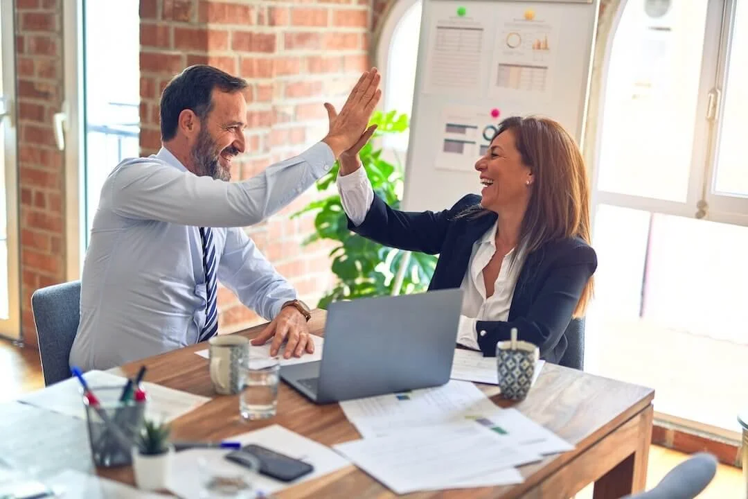 Two colleagues sitting at desk giving each other a high 5