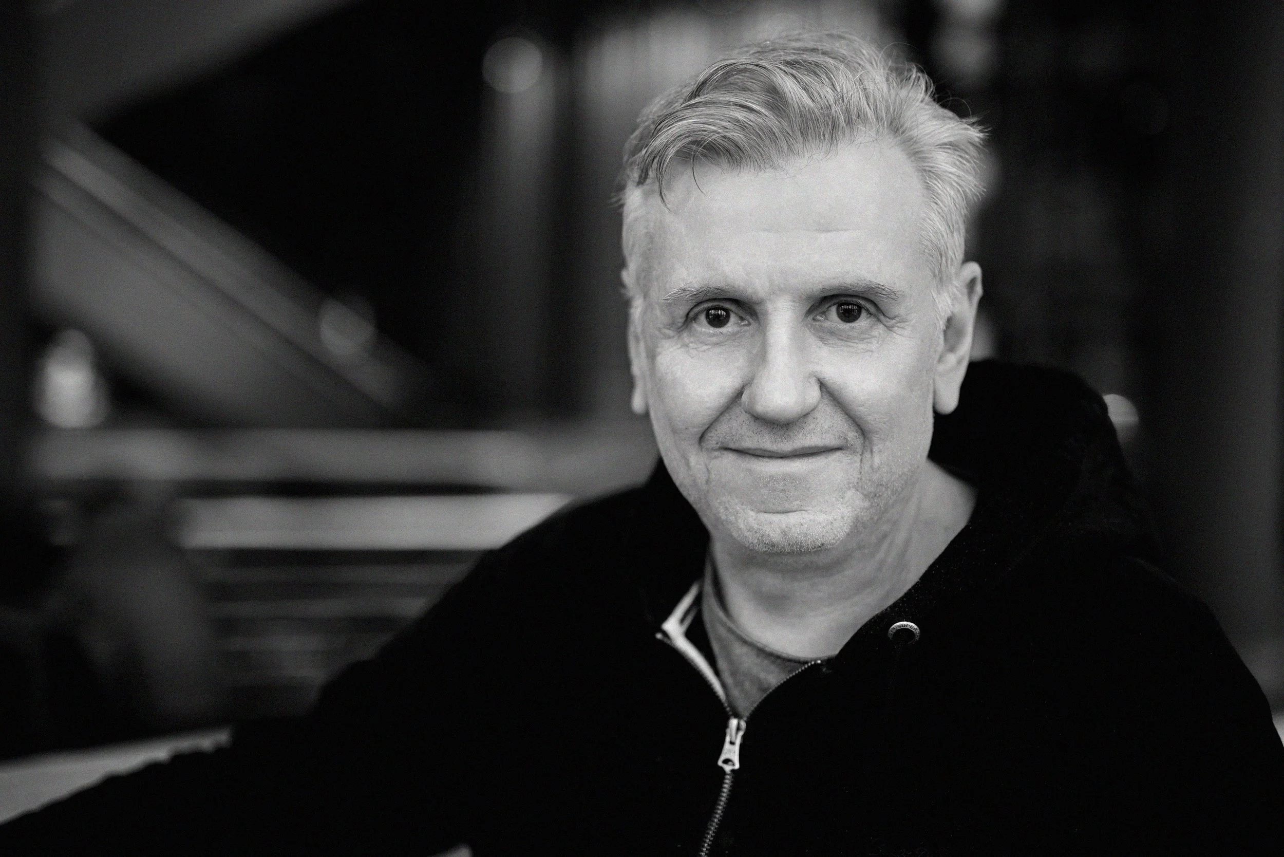 Ein schwarzer und weißer Porträt eines Mannes mit kurzen, grauen Haaren, die nach oben gestylt sind, lächelnd in die Kamera schauend, mit einem dunklen Hoodie und Hintergrund, der unscharf ist.