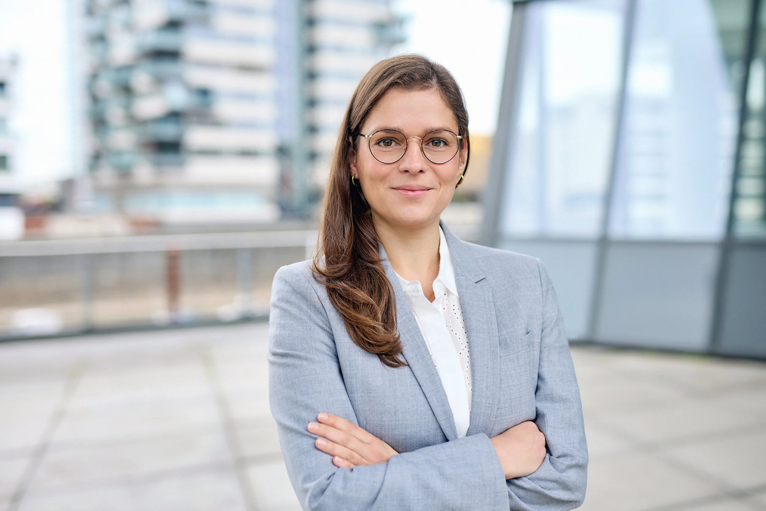 Eine junge Frau mit Brille trägt einen grauen Blazer und eine weiße Bluse, steht vor einem modernen Gebäude mit Glasfassade, lächelt leicht und hat die Arme verschränkt.