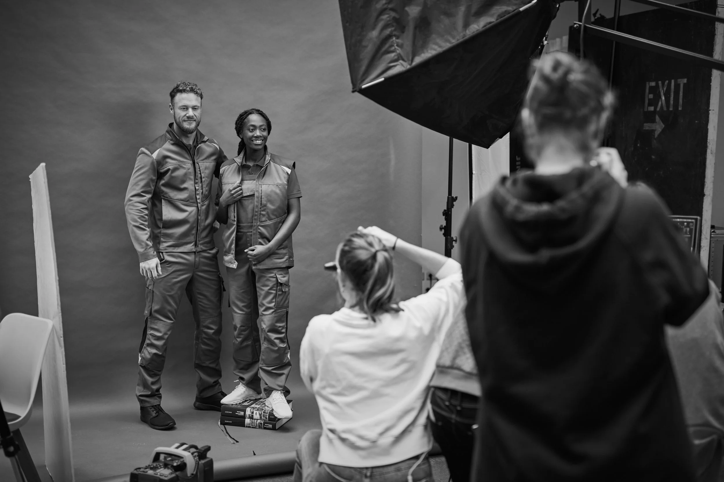 Zwei Personen posieren für ein Fotoshooting vor einem schwarzen Hintergrund, eine trägt eine Jacke mit reflektierenden Akzenten, die andere eine Weste und Cargo-Hose. Es wird von zwei Fotografen fotografiert, einer sitzt auf einem Bücherstapel, der andere steht. Professionelle Studiosetup mit Beleuchtung und Ausrüstung sichtbar.