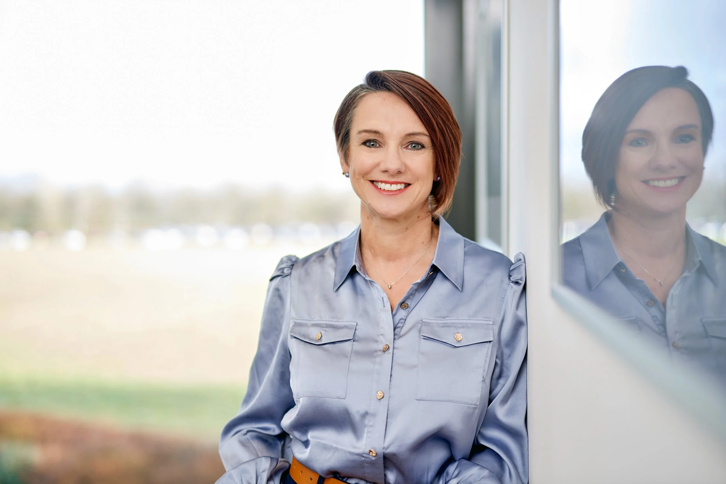 Eine Frau mit kurzen braunen Haaren und einem blauen Hemd steht vor einem Fenster und lächelt. Ihr Spiegelbild ist auf der Fensterscheibe sichtbar.