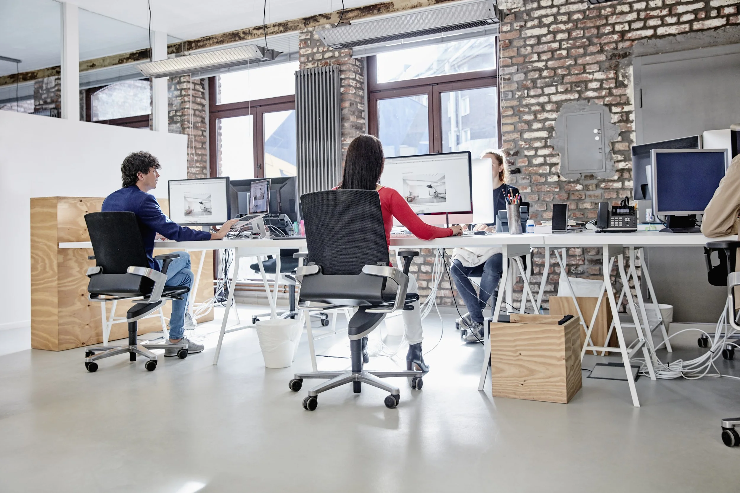 Büro mit mehreren Personen, die an Computern arbeiten, in einem Raum mit großen Fenstern, Backsteinwänden und modernen Arbeitsstationen.