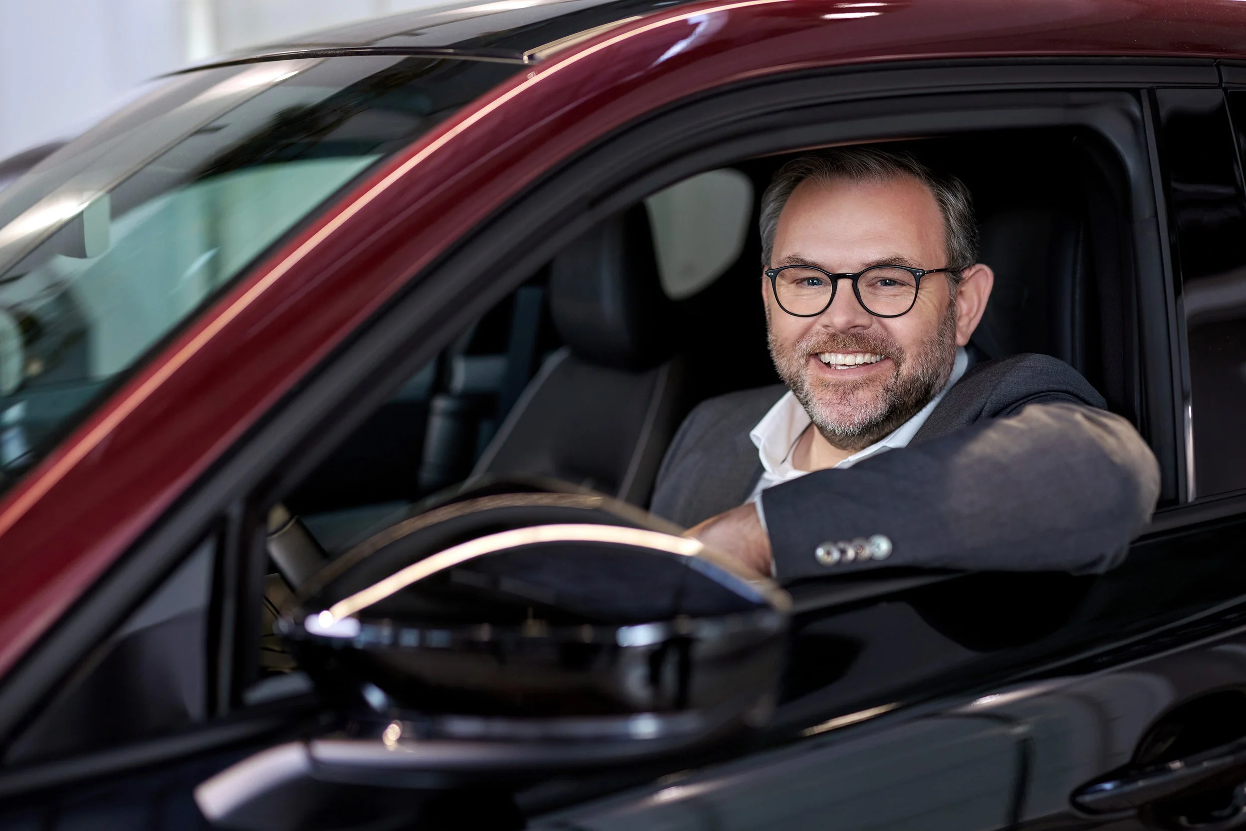 Ein lachender Mann mit Brille und grauem Bart sitzt im Auto und schaut aus dem Fenster.