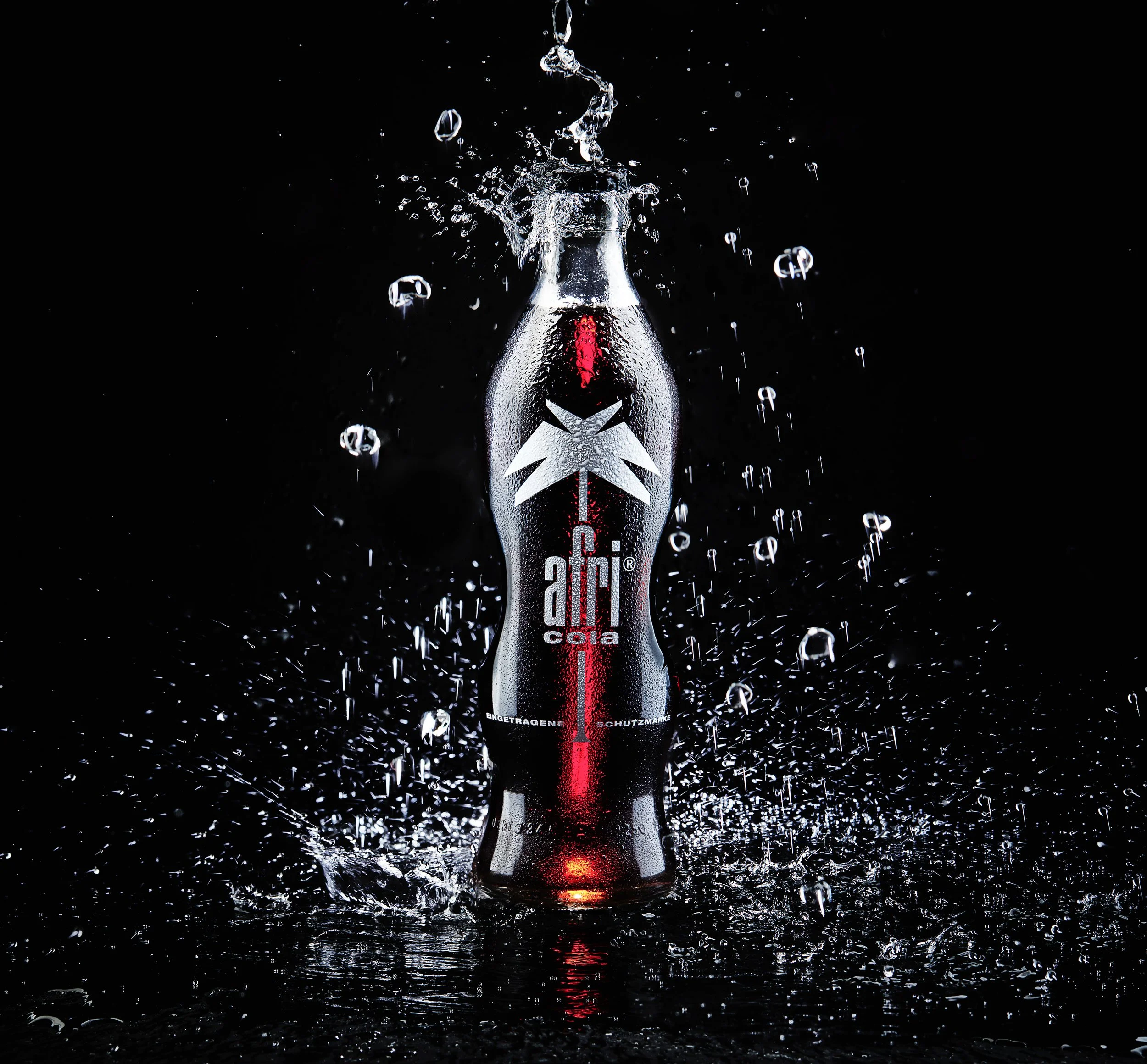 Eine aufgezogene Coca-Cola-Flasche, die in Wasser fällt, mit Wassertröpfchen und Spritzern um sie herum auf schwarzem Hintergrund.