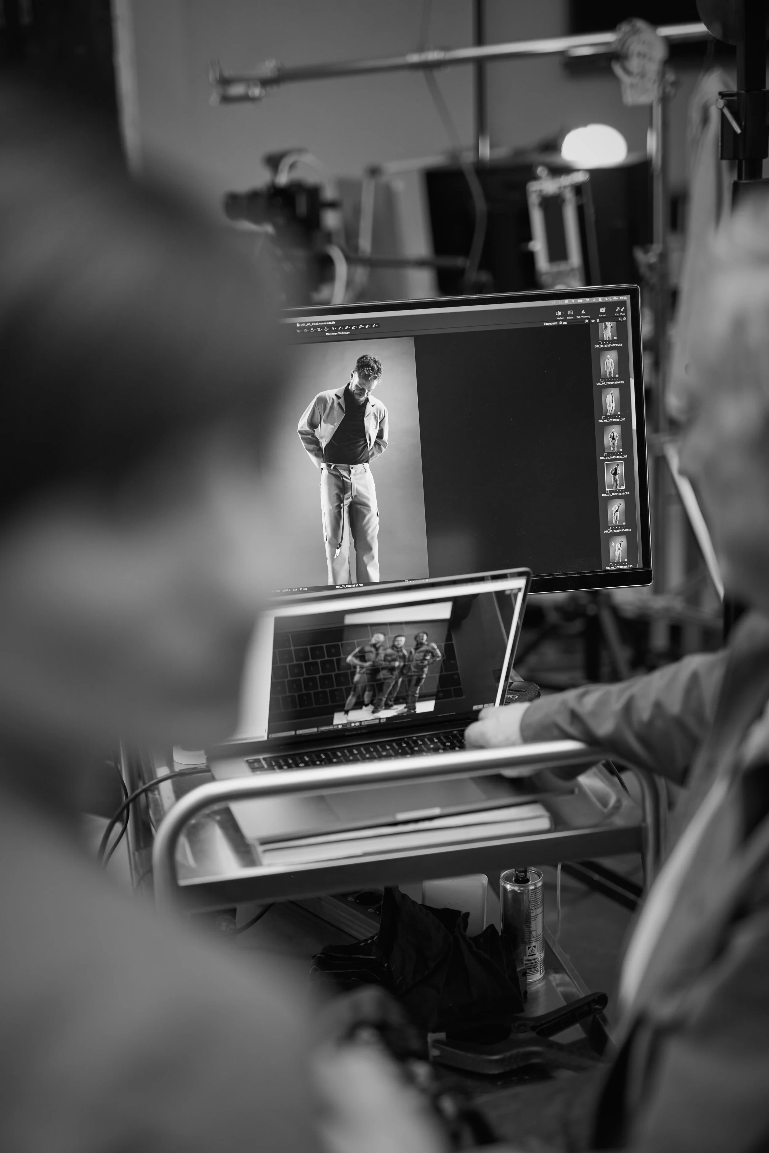Schwarz-weiß-Fotografie einer Fotoshooting-Session mit einem Model auf einem Computerbildschirm, während zwei Personen die Bilder auf einem Laptop betrachten. Im Hintergrund sind Studiolichter und technische Ausrüstung sichtbar.