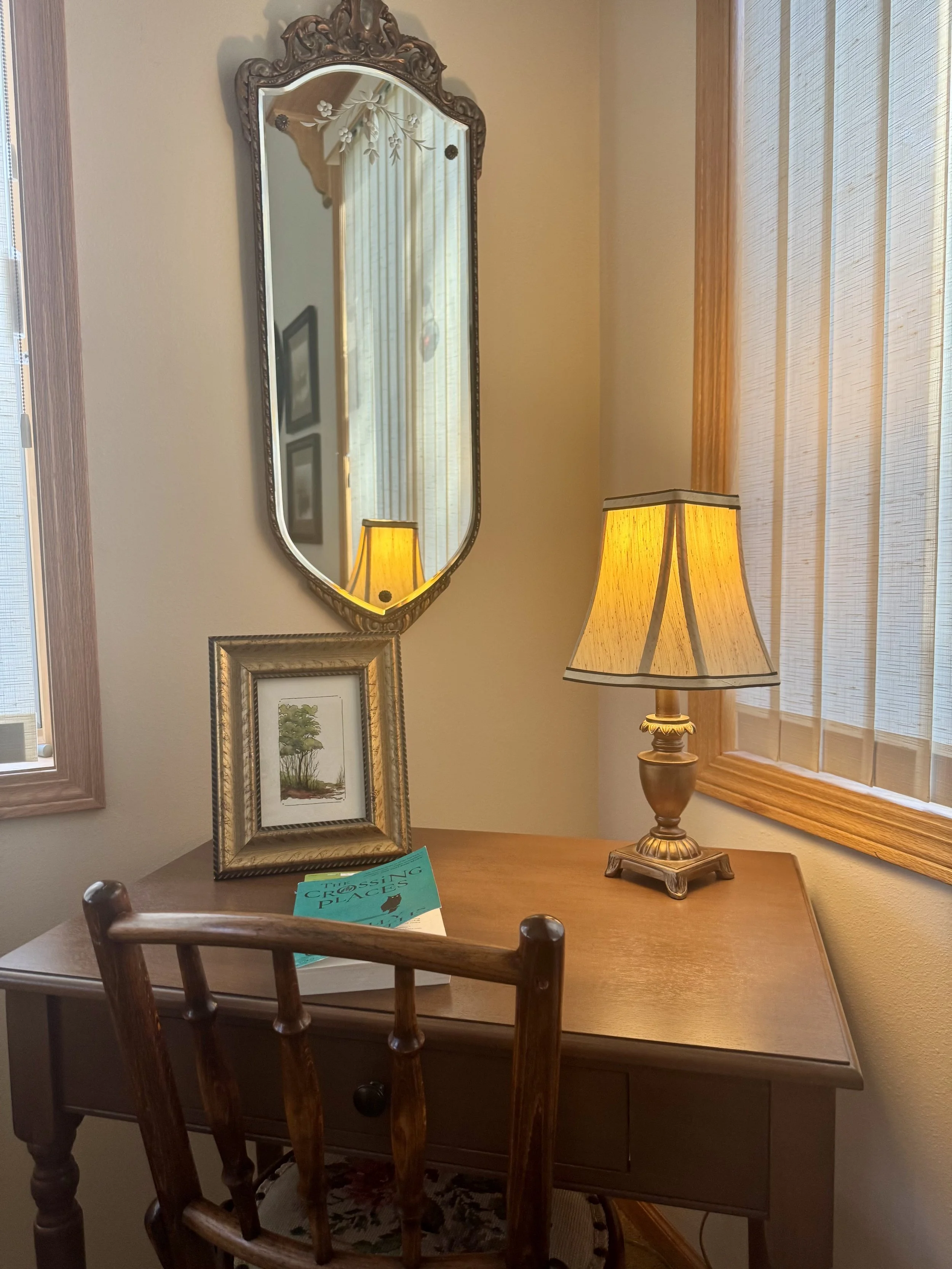 A wooden desk with a brown wooden chair in front of it, a framed painting and a turquoise book on top, a table lamp with a yellow shade, a mirror with an ornate frame on the wall, and window with wooden trim and striped blinds.