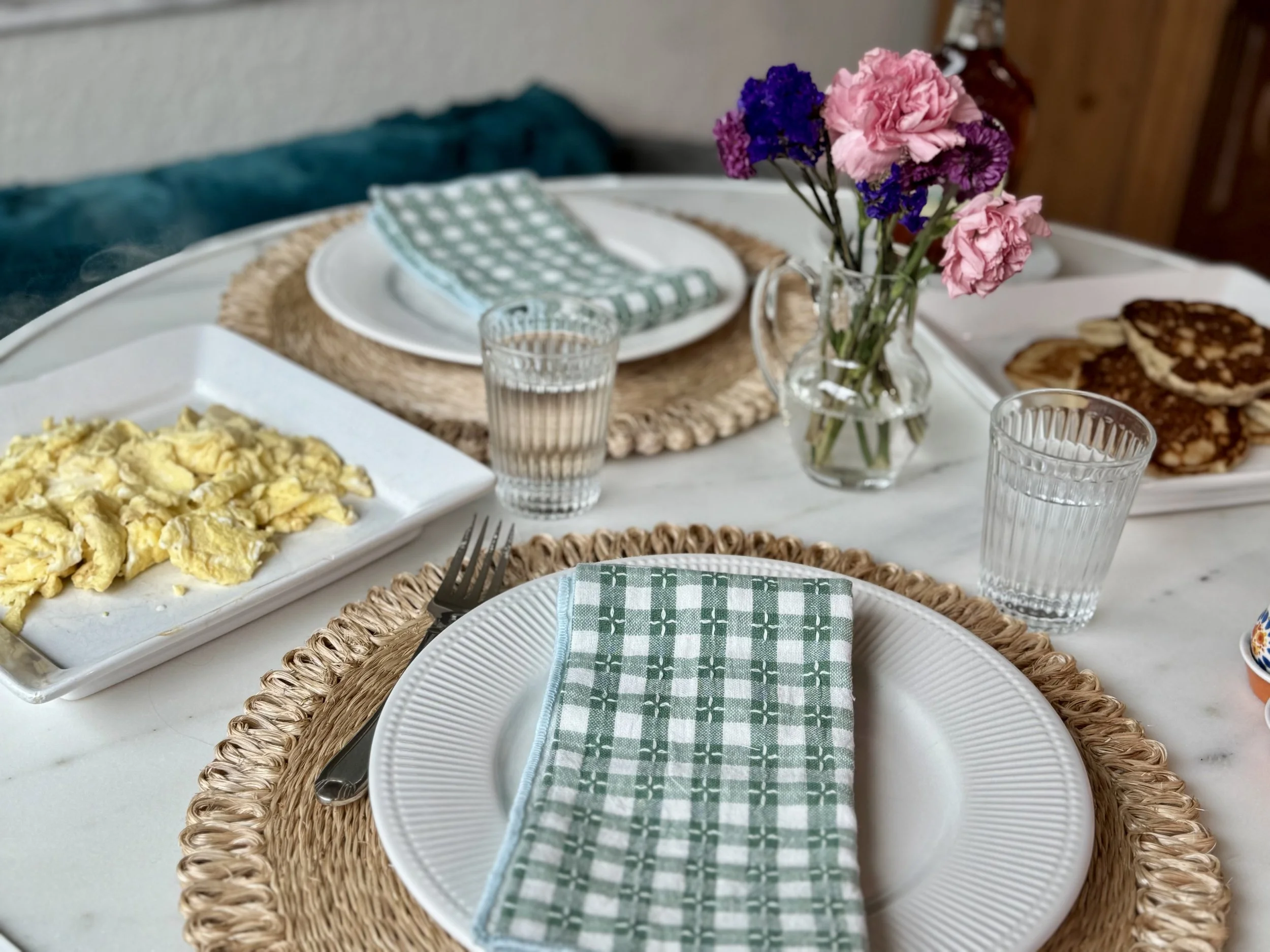 Breakfast table with scrambled eggs, pancakes, green checkered napkin, glasses of water, and a vase with pink and purple flowers.