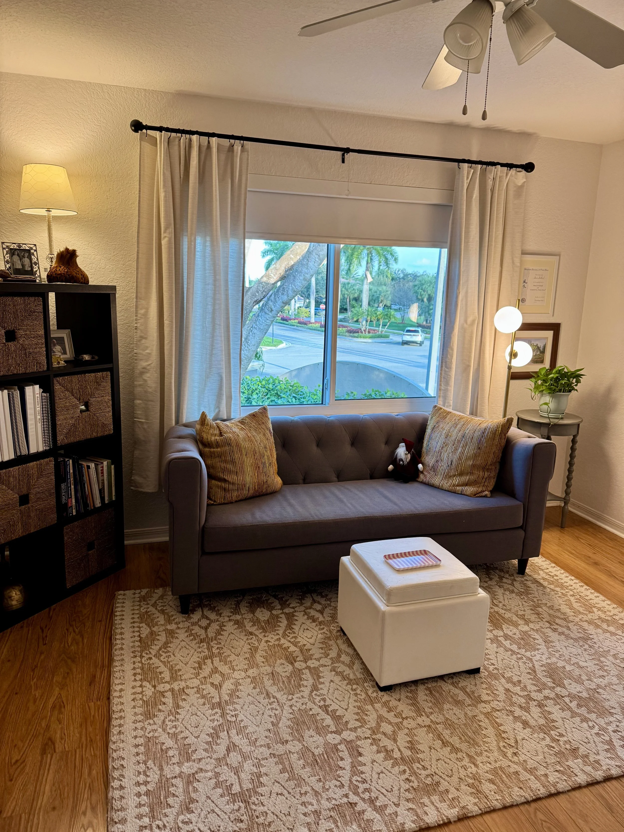 Living room with a beige couch, striped cushions, a white ottoman, a patterned rug, a black bookshelf, a table lamp, and a window with light curtains showing trees and street outside.
