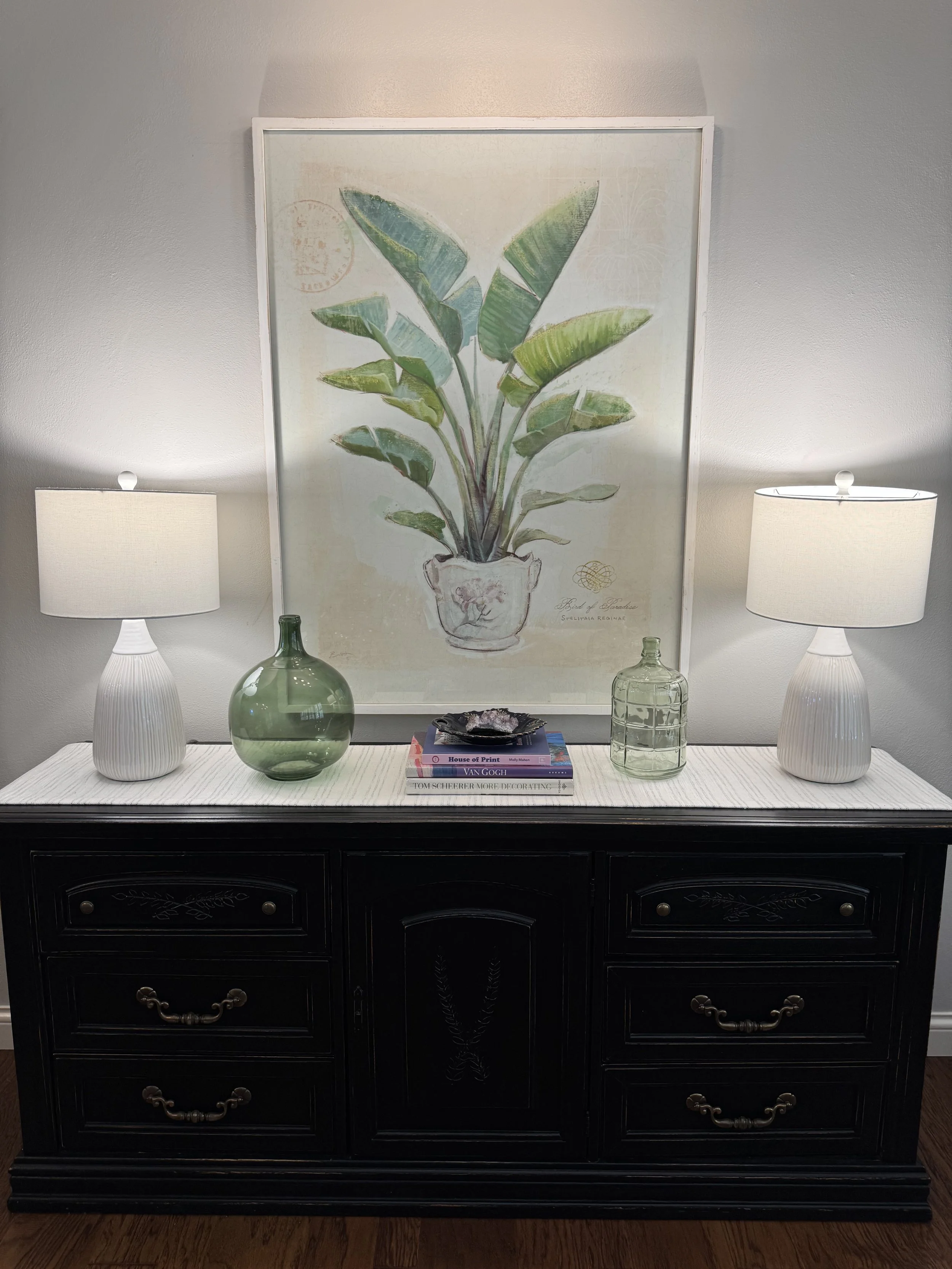 A black wooden dresser with three drawers, brass handles, and decorative carvings, topped with a white table runner, two white lamps with beige shades, a large green glass vase, a smaller glass container, a stack of books, and decorative items, with a large botanical print of a potted plant hanging on the wall behind.