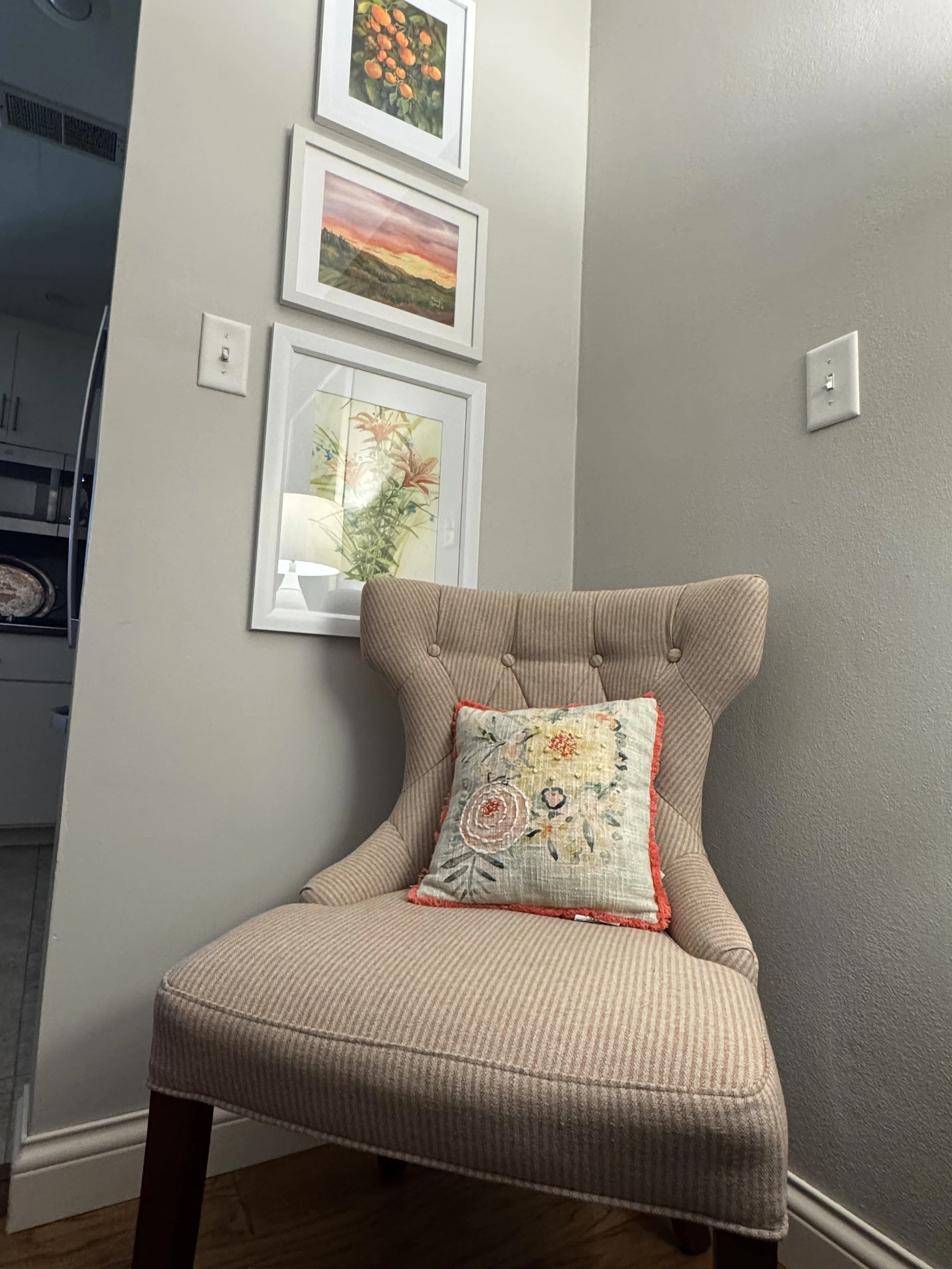 A beige upholstered armchair with a floral pillow placed on it, positioned against a wall with three framed landscape and floral artworks. The chair is near a corner in a room with hardwood flooring.