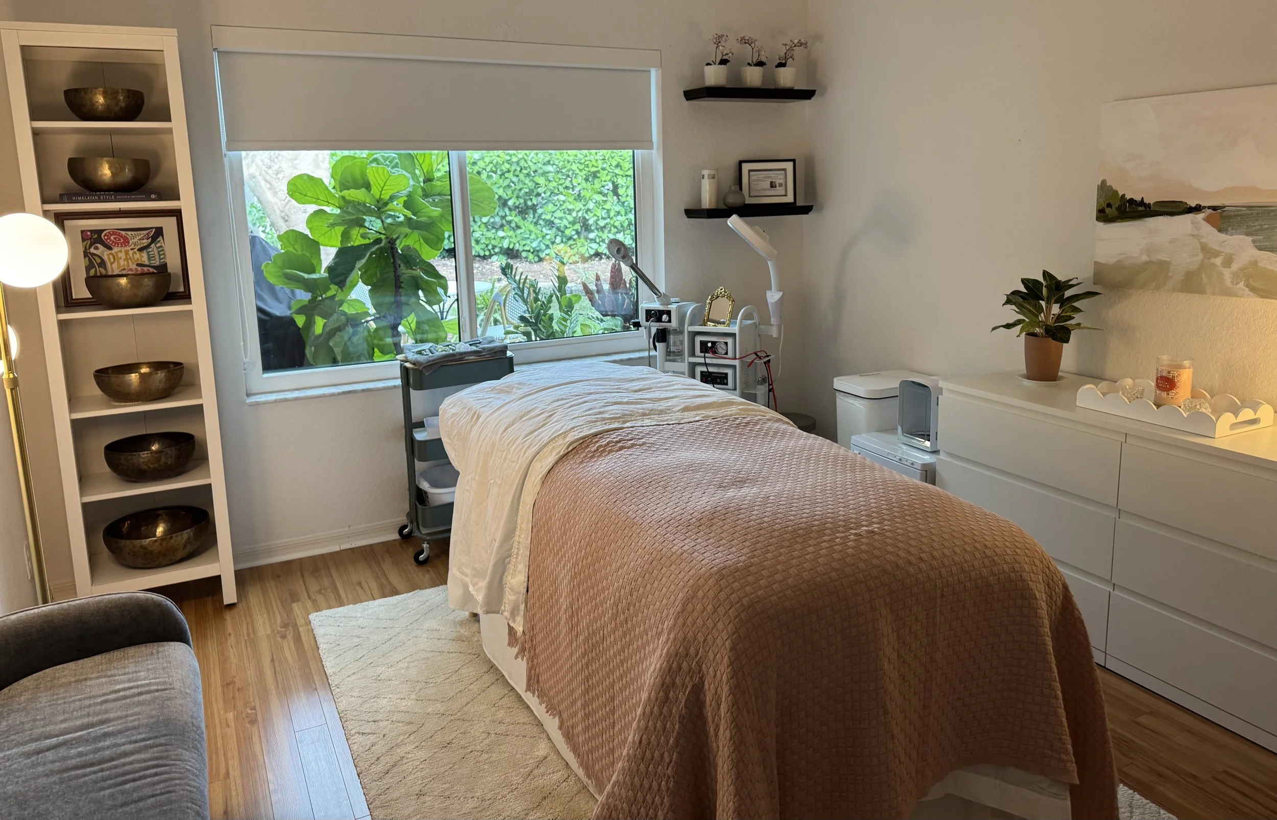 A bedroom with a bed, decorative shelves, plants, a window with greenery outside, and various decorative items.