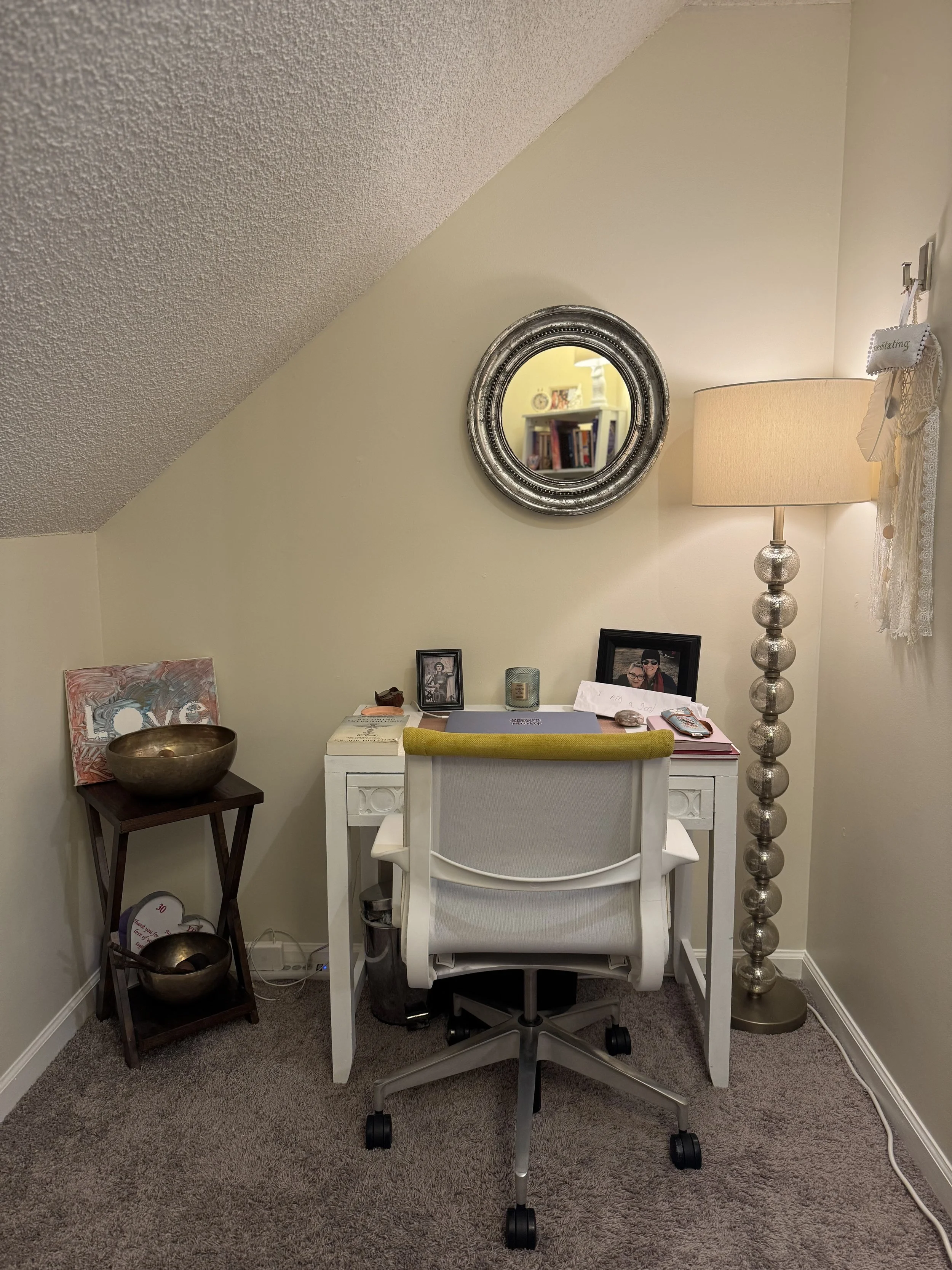 A white desk with a yellow chair, a black photo frame, a lamp, and various decorative items, against a beige wall with a circular silver mirror. To the right, a tall silver floor lamp with glass orbs. A small table with metal bowls and framed photos on the left, and a power strip on the floor. Carpeted floor.