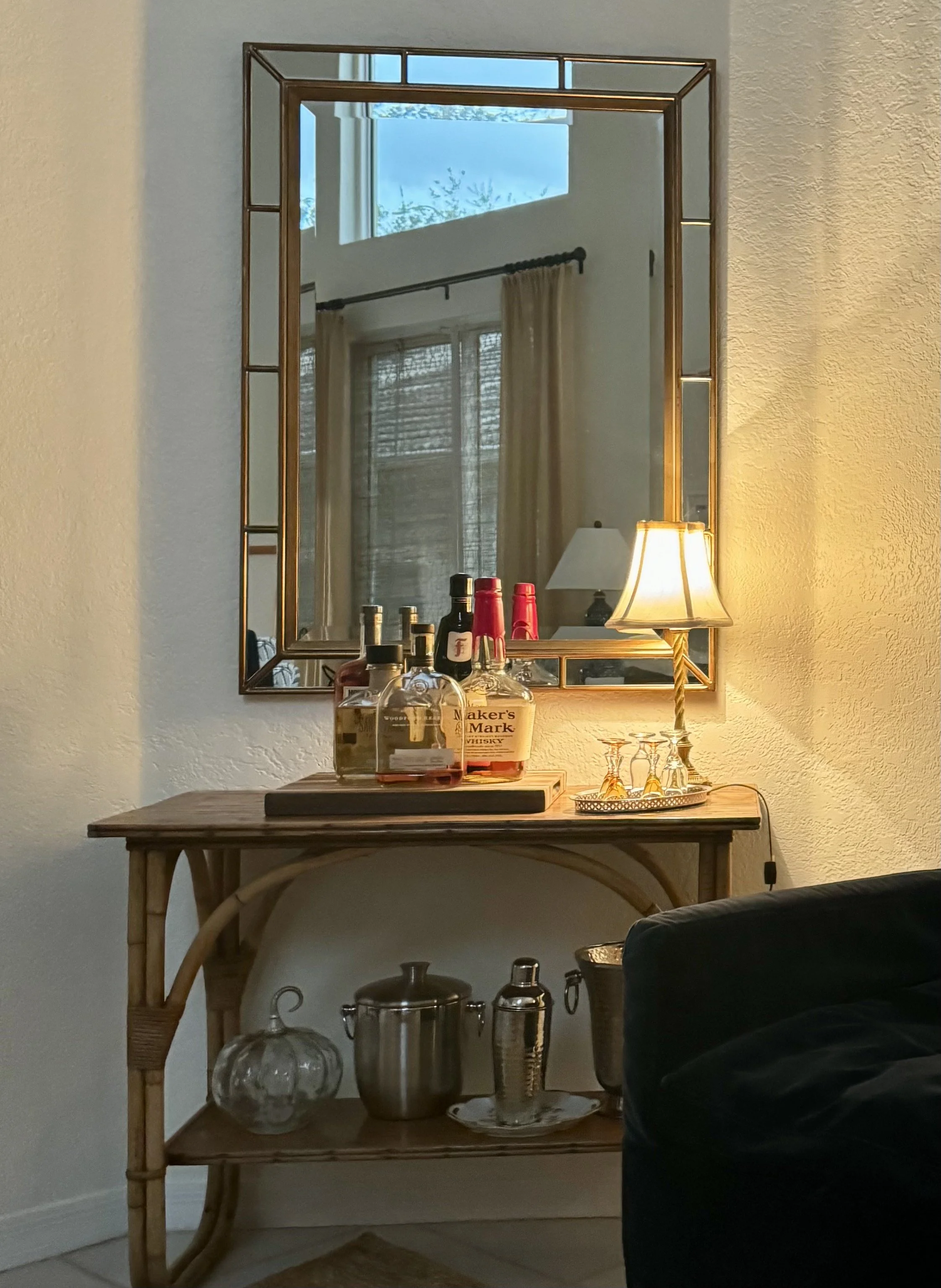 A wooden side table with a few bottles of alcohol on top, positioned in front of a large mirror with a gold-colored frame. The mirror reflects a window with closed blinds, curtains, and part of the ceiling. A small lamp is on the right side of the mirror. The lower shelf of the side table holds various glassware and barware, including a large glass pitcher, a metal cocktail shaker, and a gold-colored vessel, with a tray of glasses on the right.