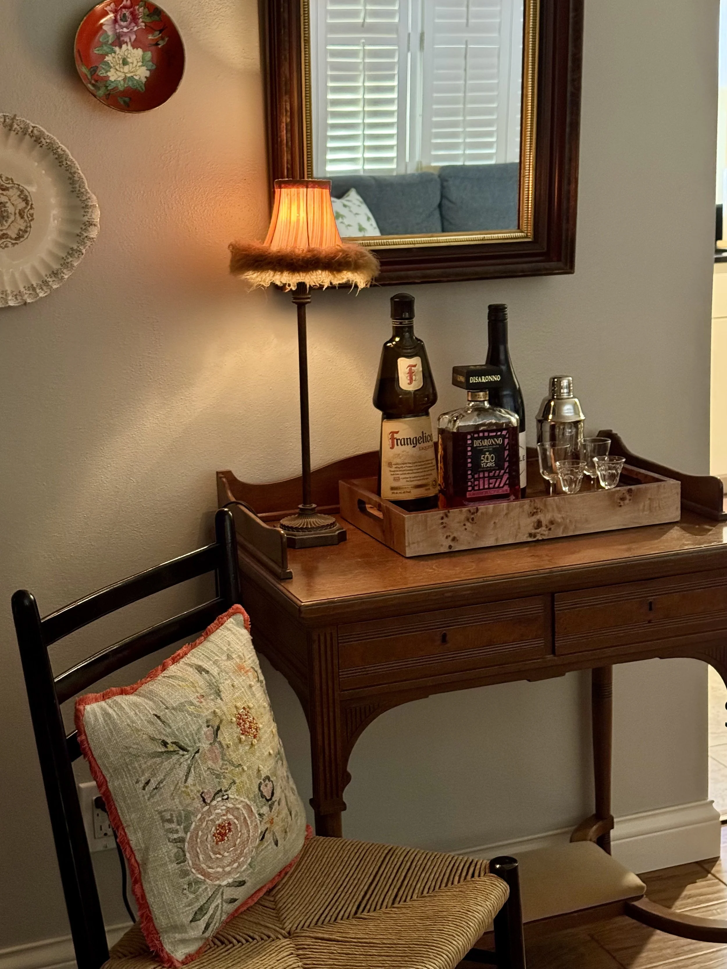 A wooden side table with a tray holding bottles of liquor and shot glasses, beside a black dining chair with a decorative needlepoint pillow, a table lamp, and a mirror on the wall reflecting a blue sofa with a pillow and window shutters.