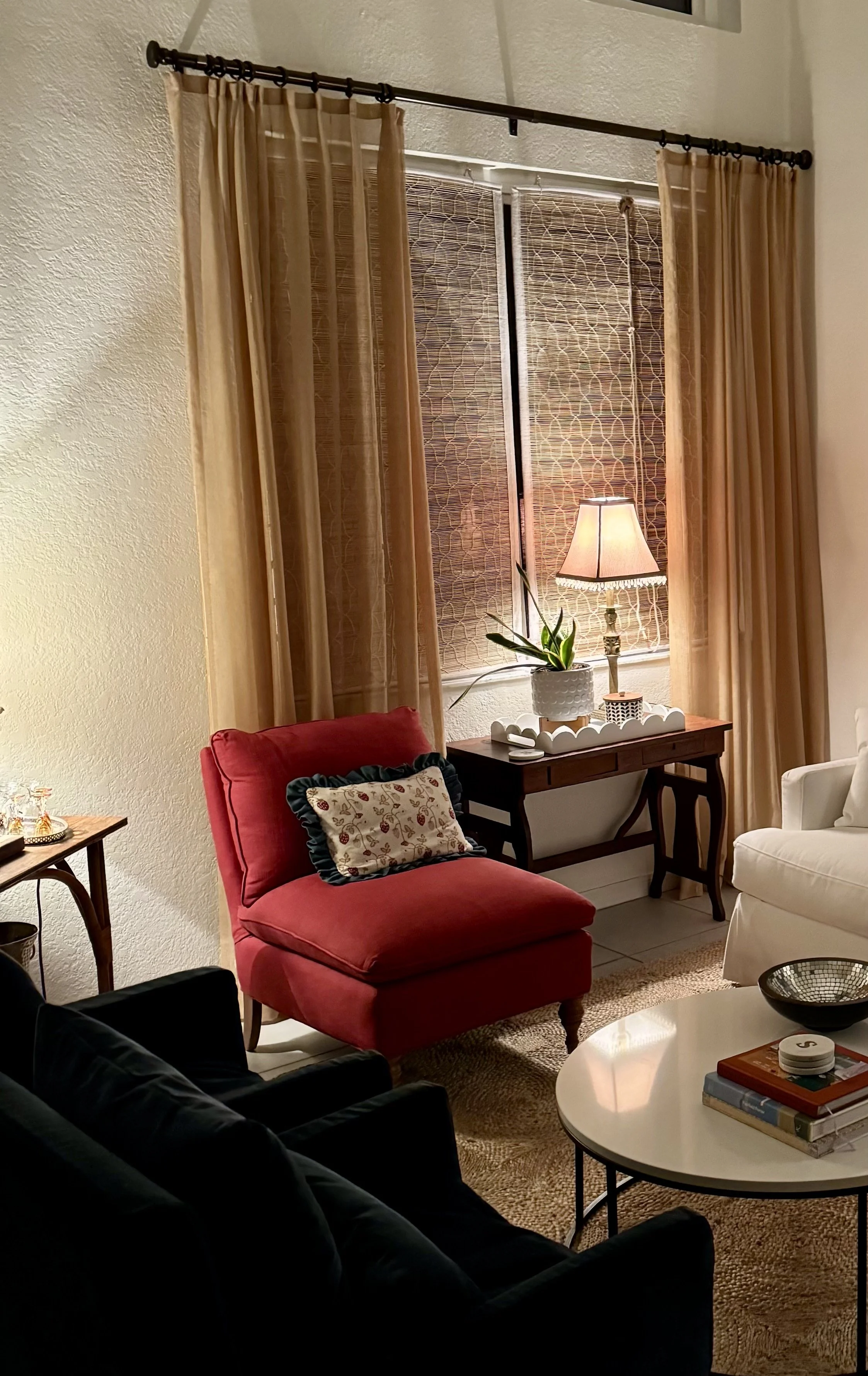 Living room with red armchair, white armchair, black sofa, round white coffee table, table with books, tray, ball, lamp on side table, window with bamboo shades, beige curtains, plant, decorative candle, and picture frame.
