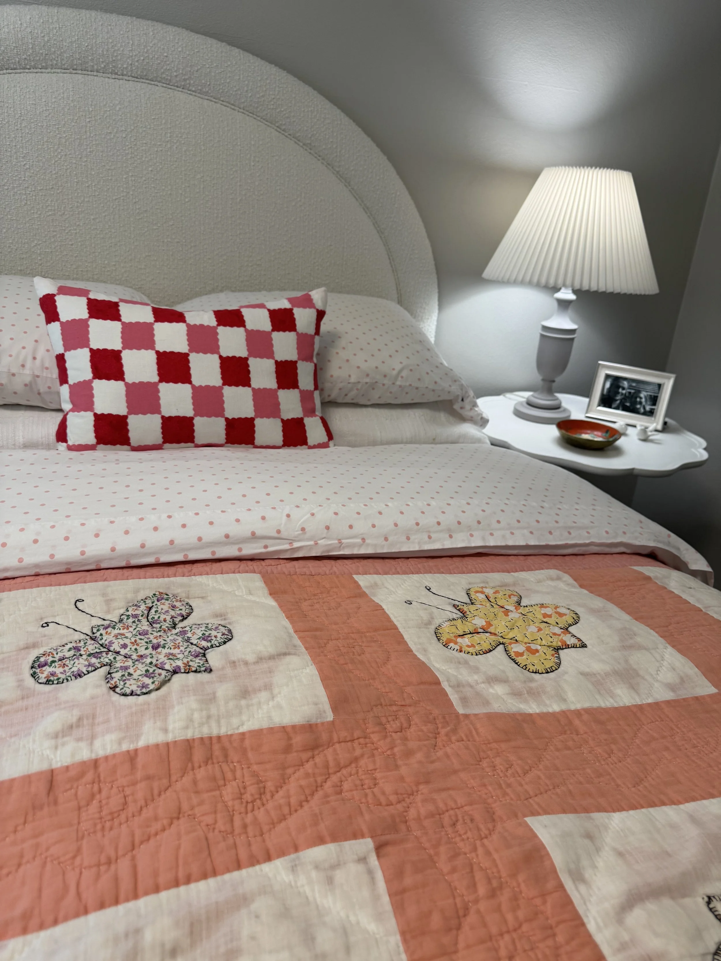 A bed with a pink and white polka dot bedsheet, a red and white checkered pillow, a quilt with butterfly embroidery, a bedside table with a framed photo, a bowl, and a white lamp
