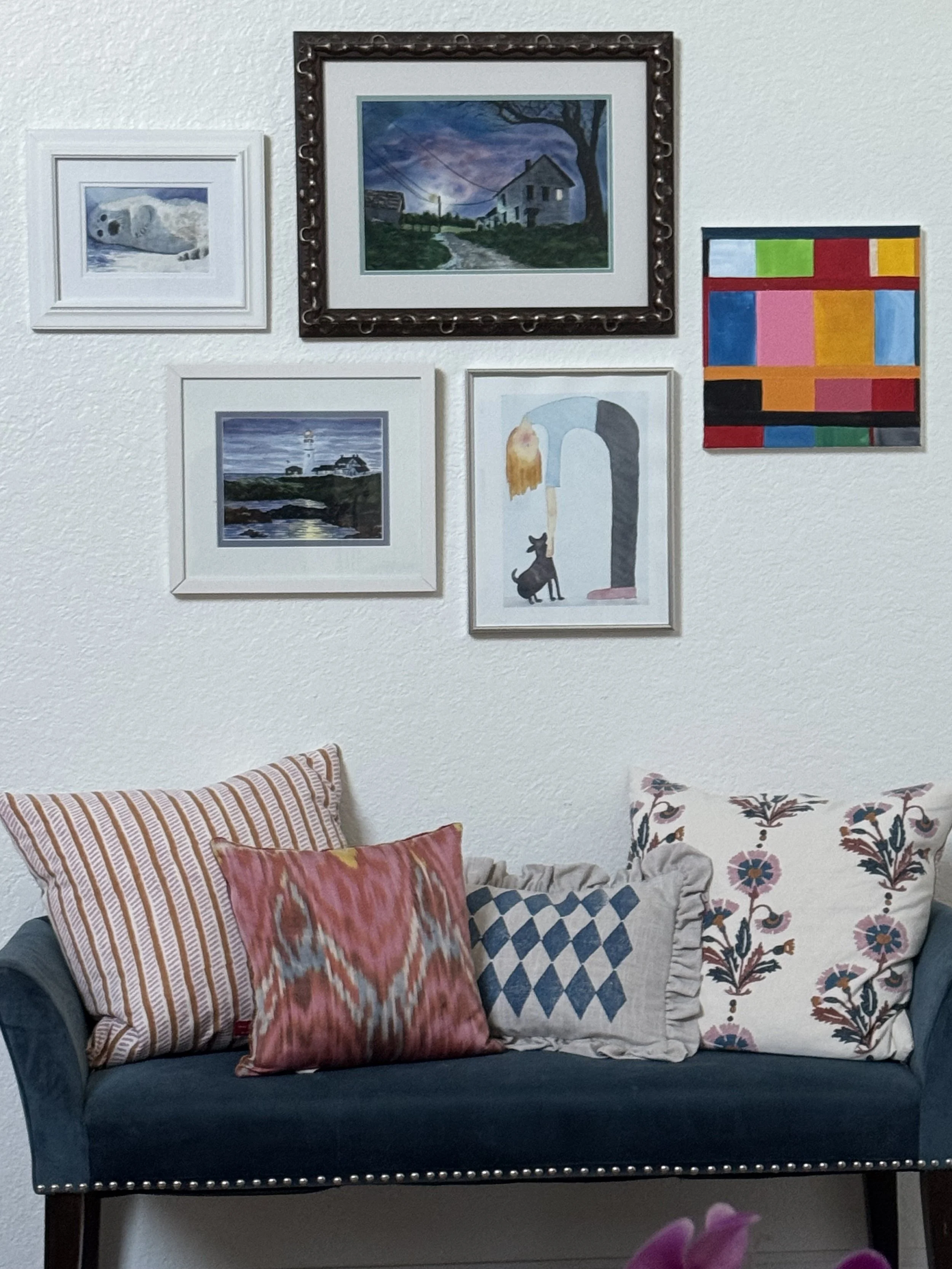 A dark blue sofa with multiple decorative pillows, including pink, orange, black, blue, and floral patterns, in a living room with wall art above.