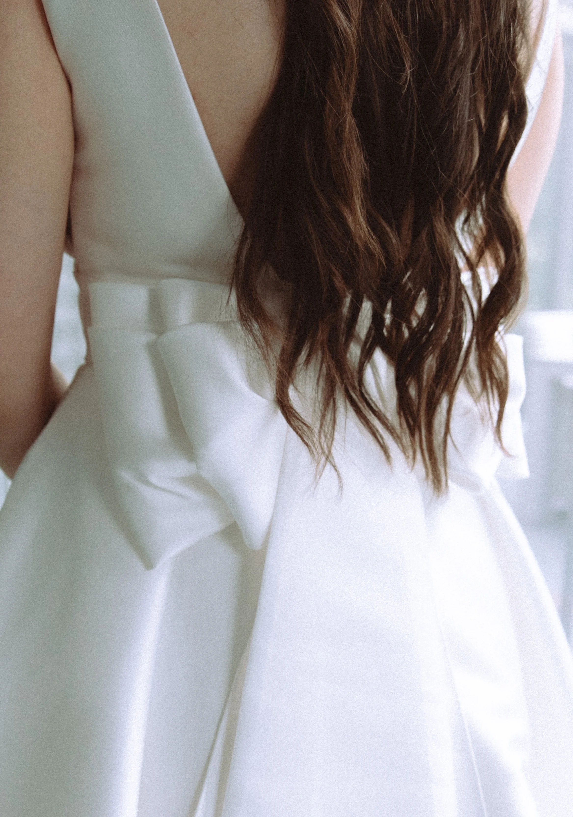 Close-up of a woman wearing a white sleeveless dress with a bow detail at the waist, long wavy brown hair.