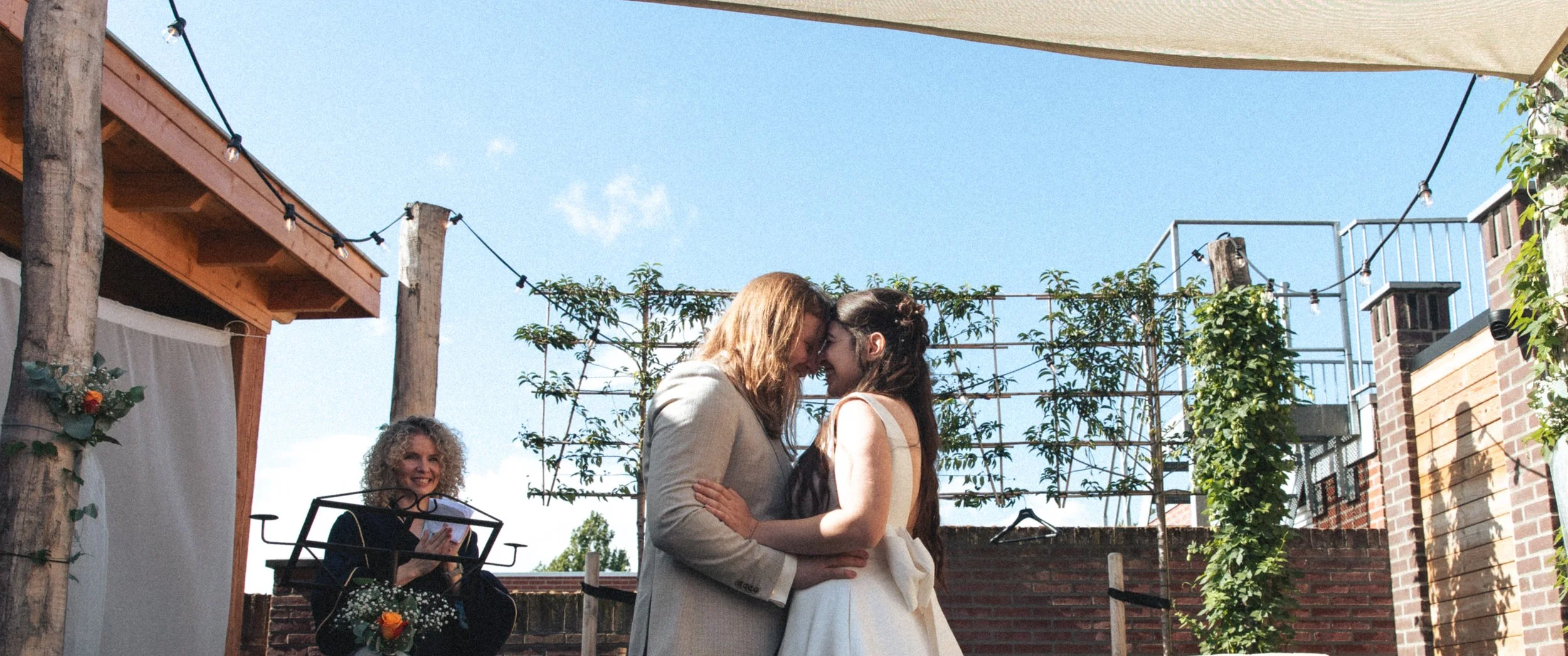 Couple hugging at their wedding ceremony outdoors, with a woman in a black dress holding flowers smiling in the background, under a canopy and string lights.