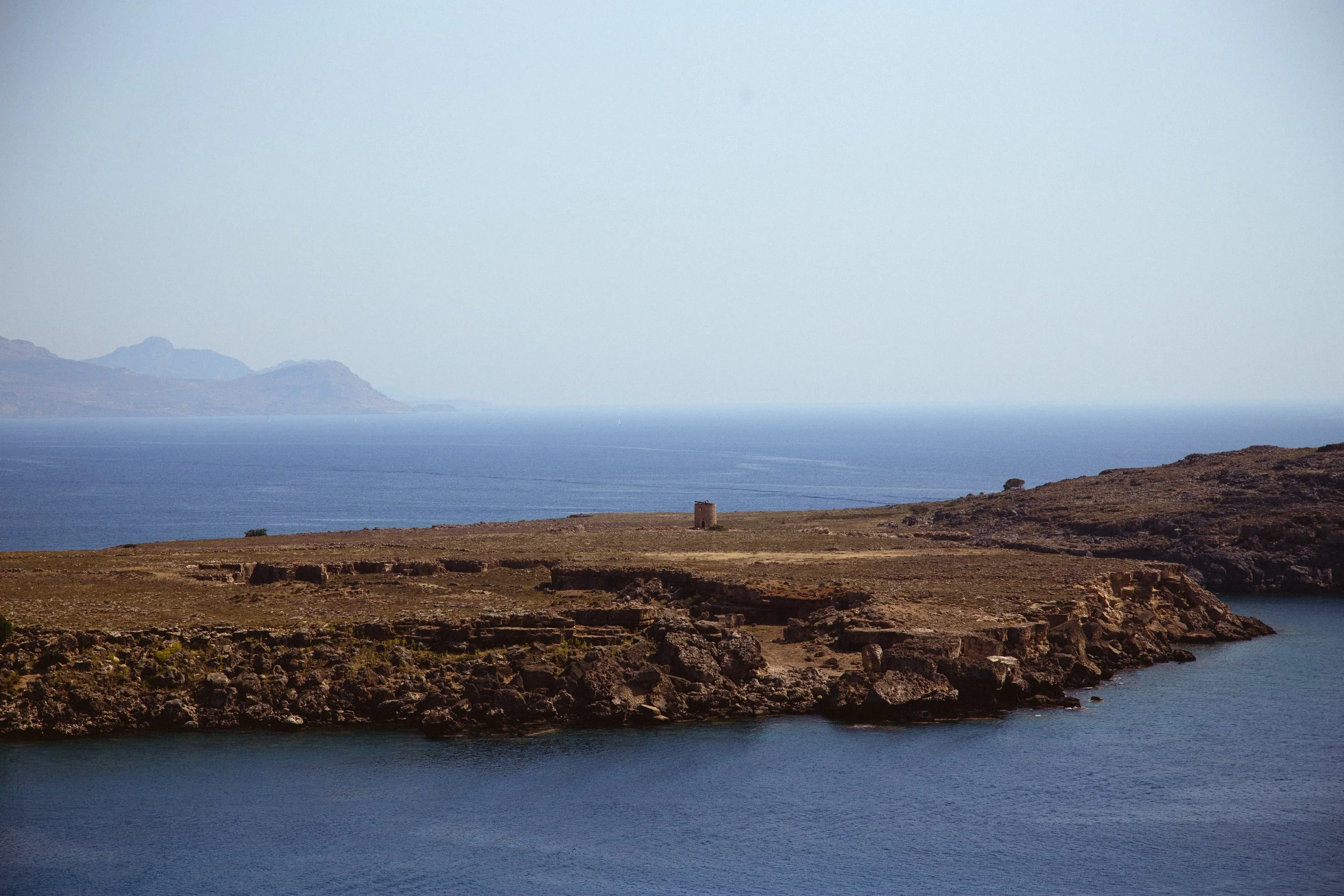 A coastal landscape featuring rocky terrain, calm water, and a structure on the land with mountains in the background.