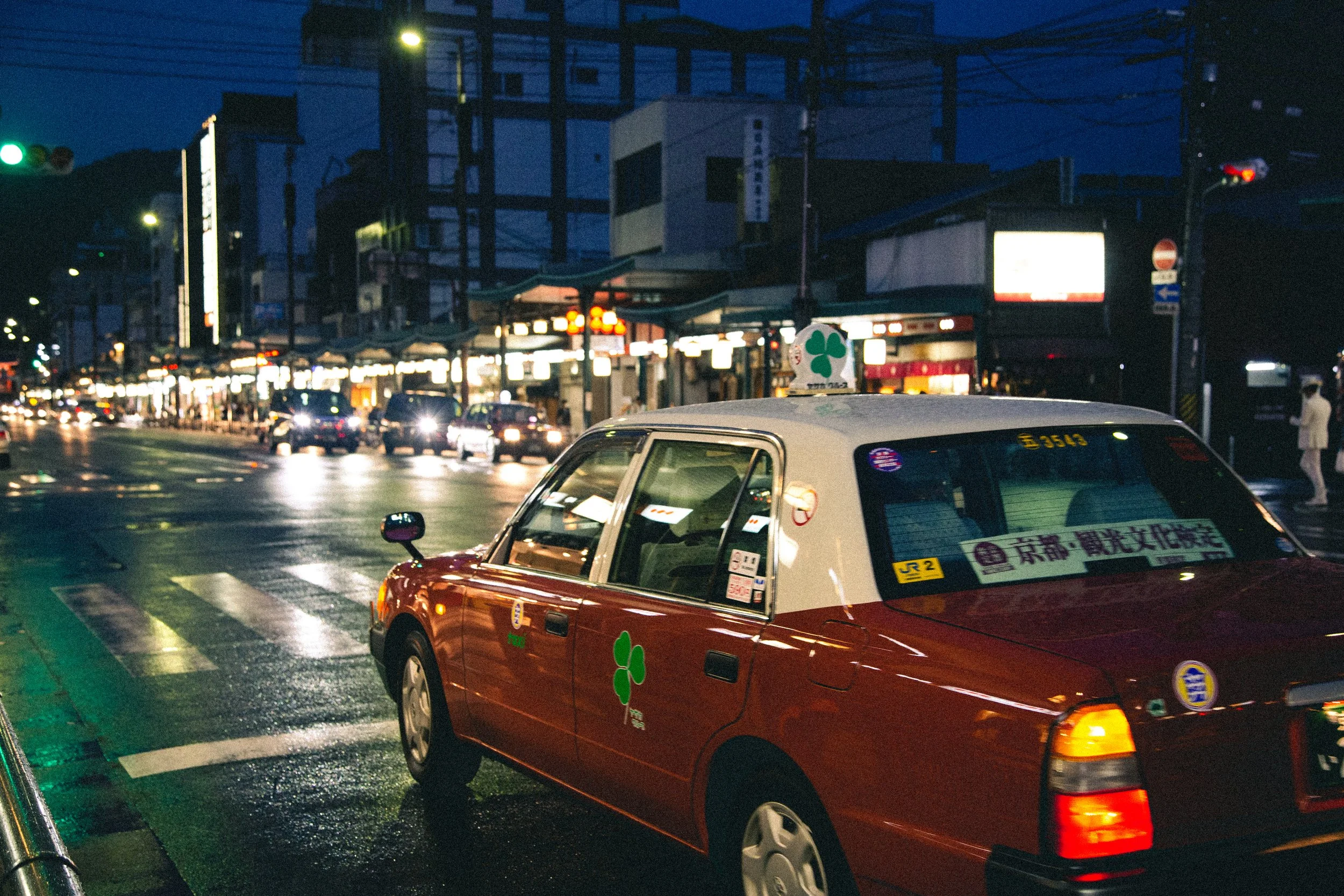 Nighttime city street with a red taxi in the foreground, amid other cars, illuminated signs, and buildings in the background.