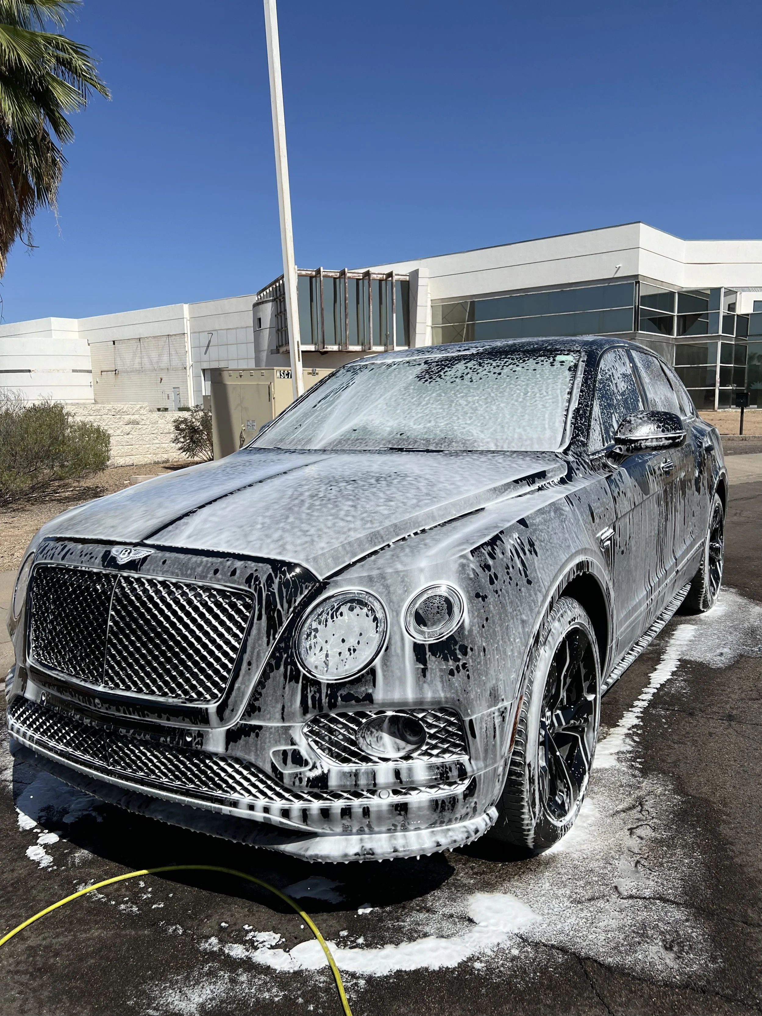 A black luxury SUV being washed with soap and foam, parked outdoors with a modern building and palm trees in the background.