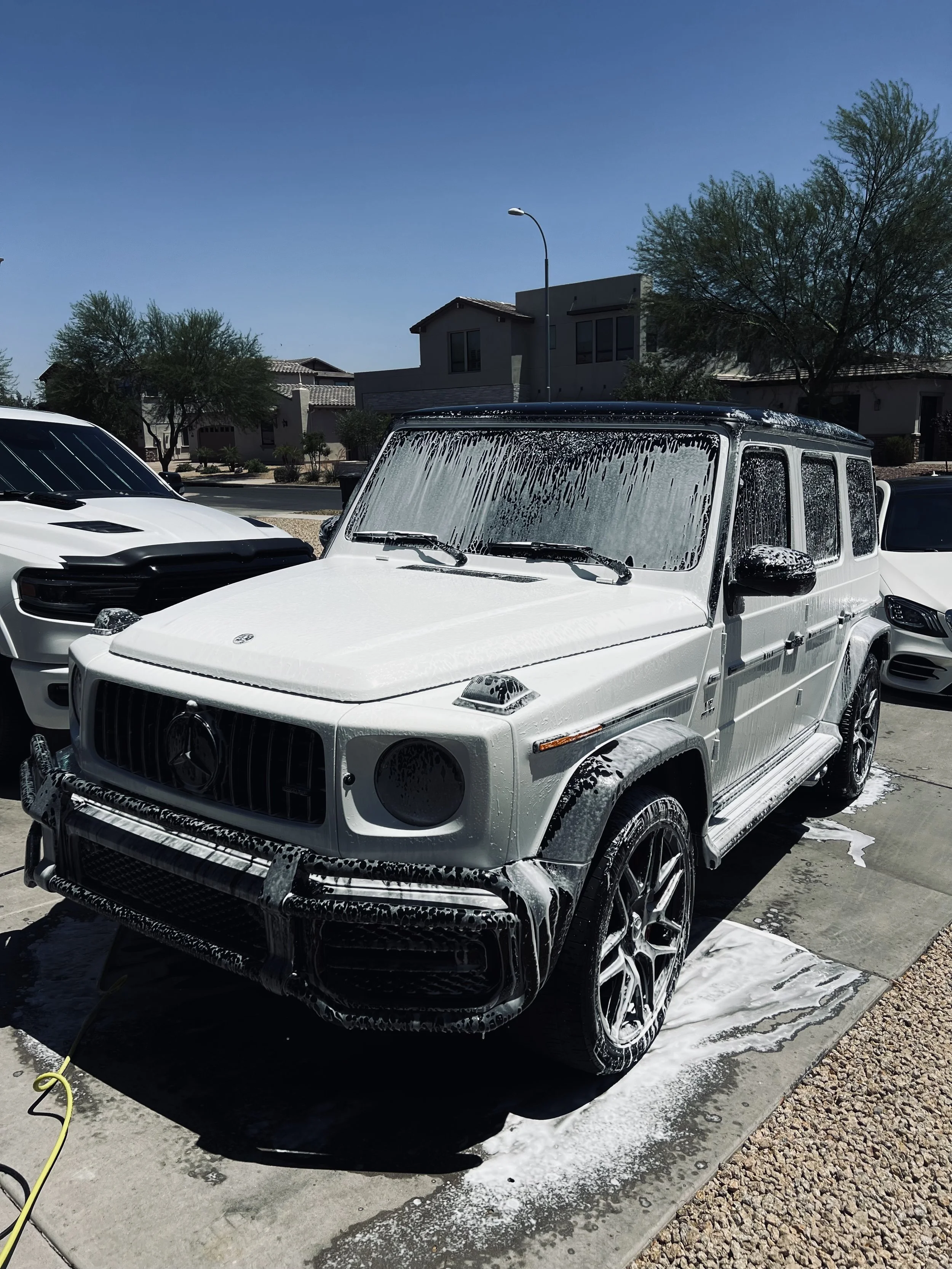 White Mercedes-Benz G-Class SUV being washed, foam and soap on the vehicle, in a residential driveway on a sunny day.