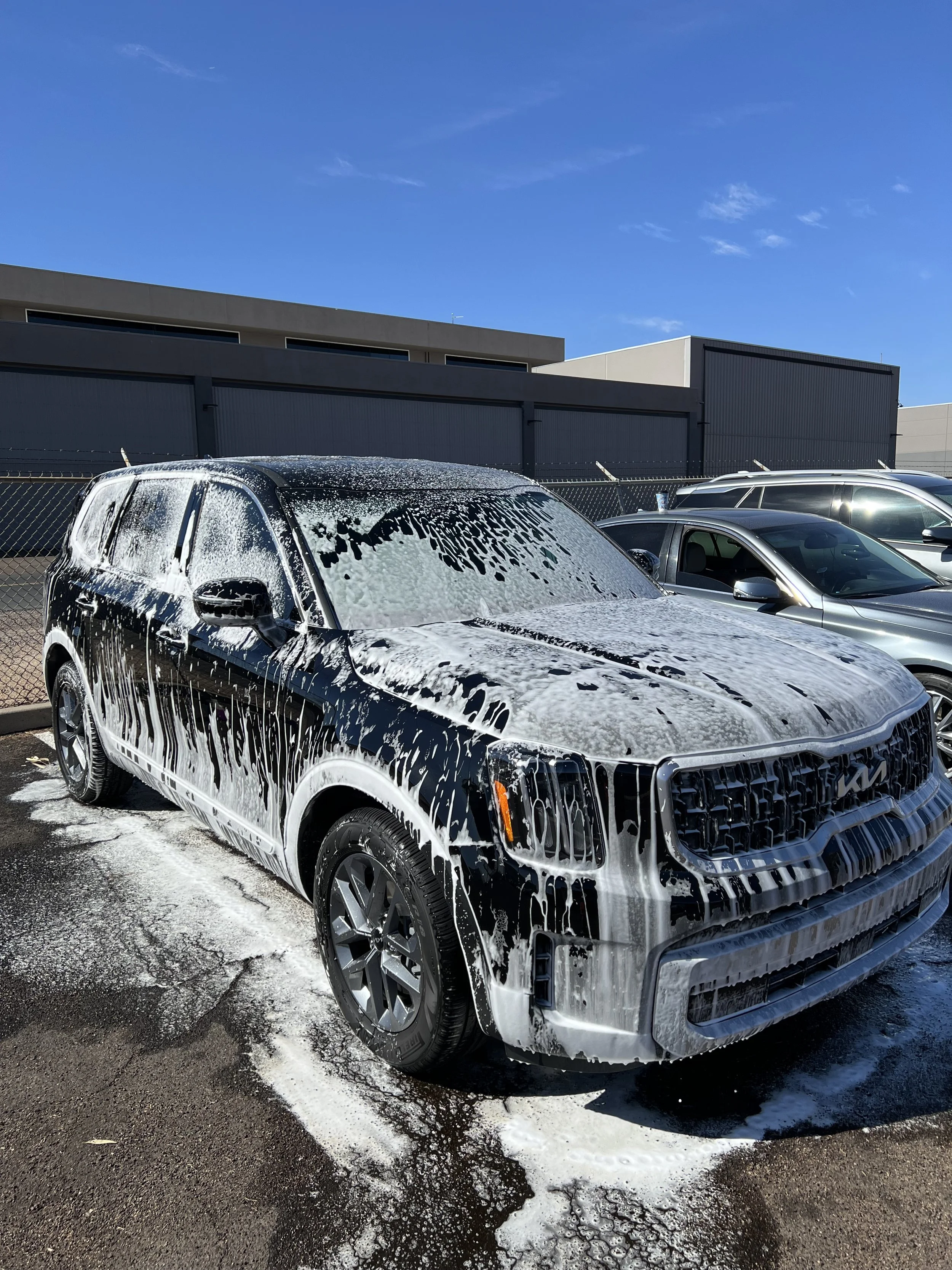 Black SUV covered in soap suds during a car wash, with blue sky and other vehicles in the background.