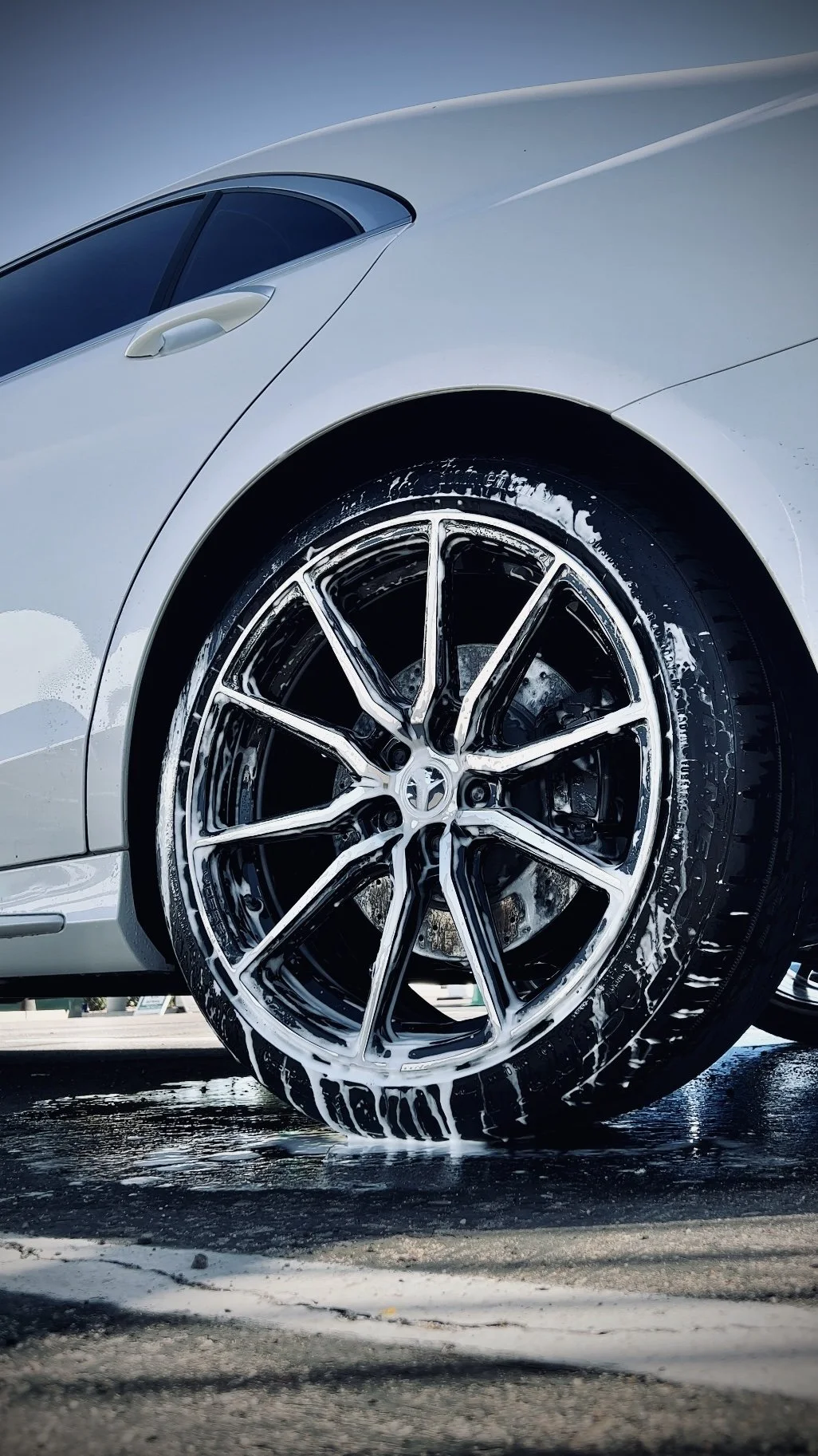 Close-up of a silver sports car wheel being washed with soap, with water and soap suds on the ground.