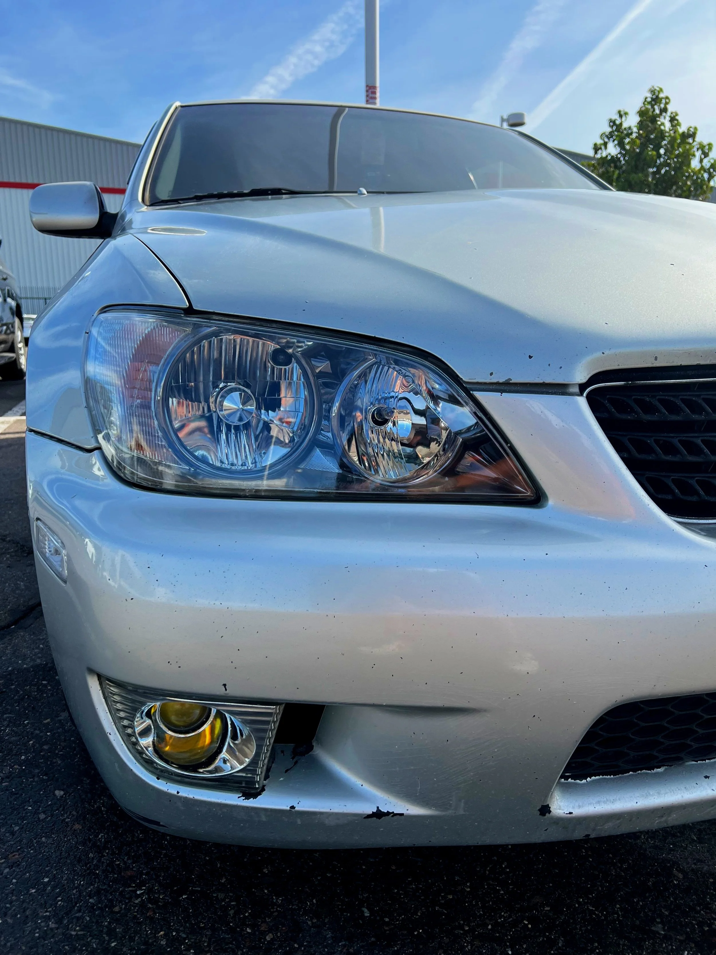 Front view of a silver sedan with a damaged lower front bumper and a keyhole for the hood open, parked outdoors on asphalt with a building and trees in the background.
