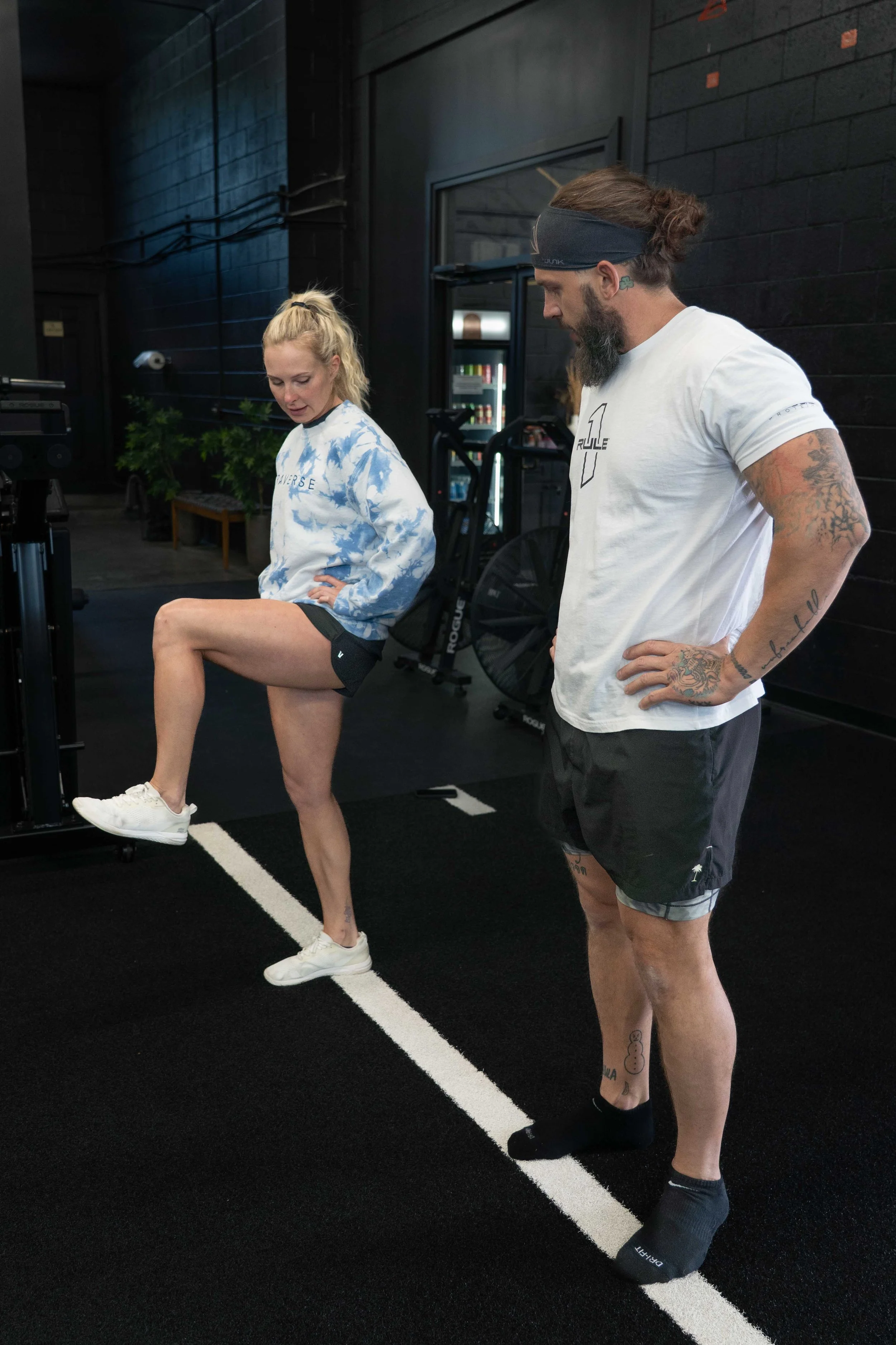 A woman in athletic wear is performing a leg raise as a man watches and coaches her in a gym with black walls and fitness equipment.