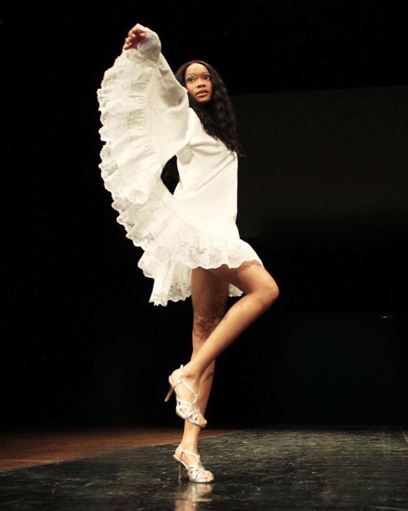 A woman in a white dress with ruffled sleeves and hem, standing on a stage with one leg raised, wearing high heel shoes, against a black background.