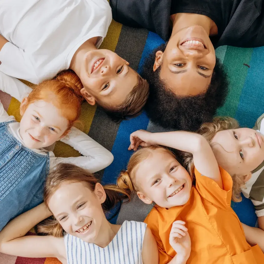 A bunch of kids and one adult laying on the ground in a circle looking and smiling at the camera.