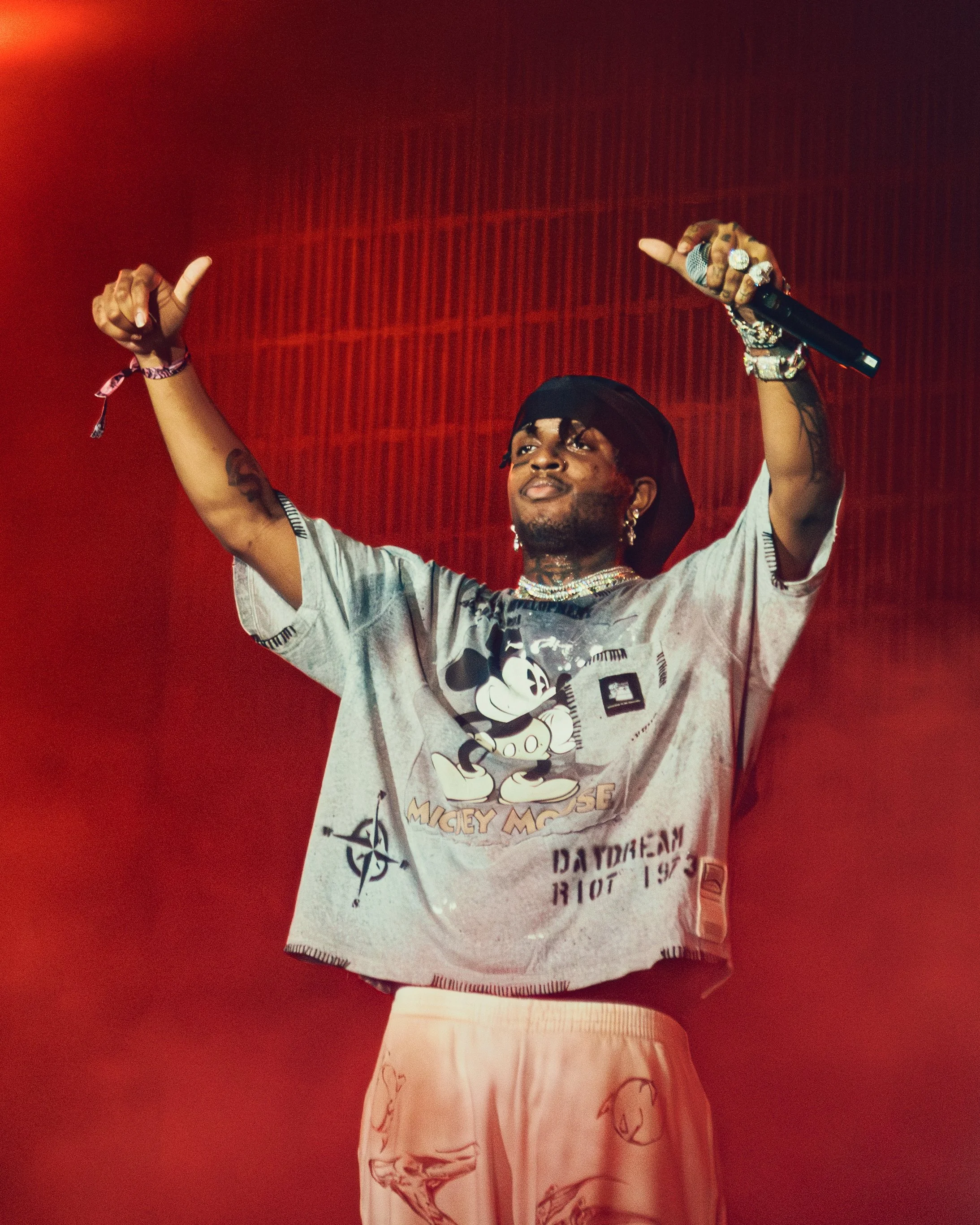A man standing on stage with his arms raised, holding a microphone, wearing a Mickey Mouse t-shirt, pink shorts, and jewelry, against a red background.