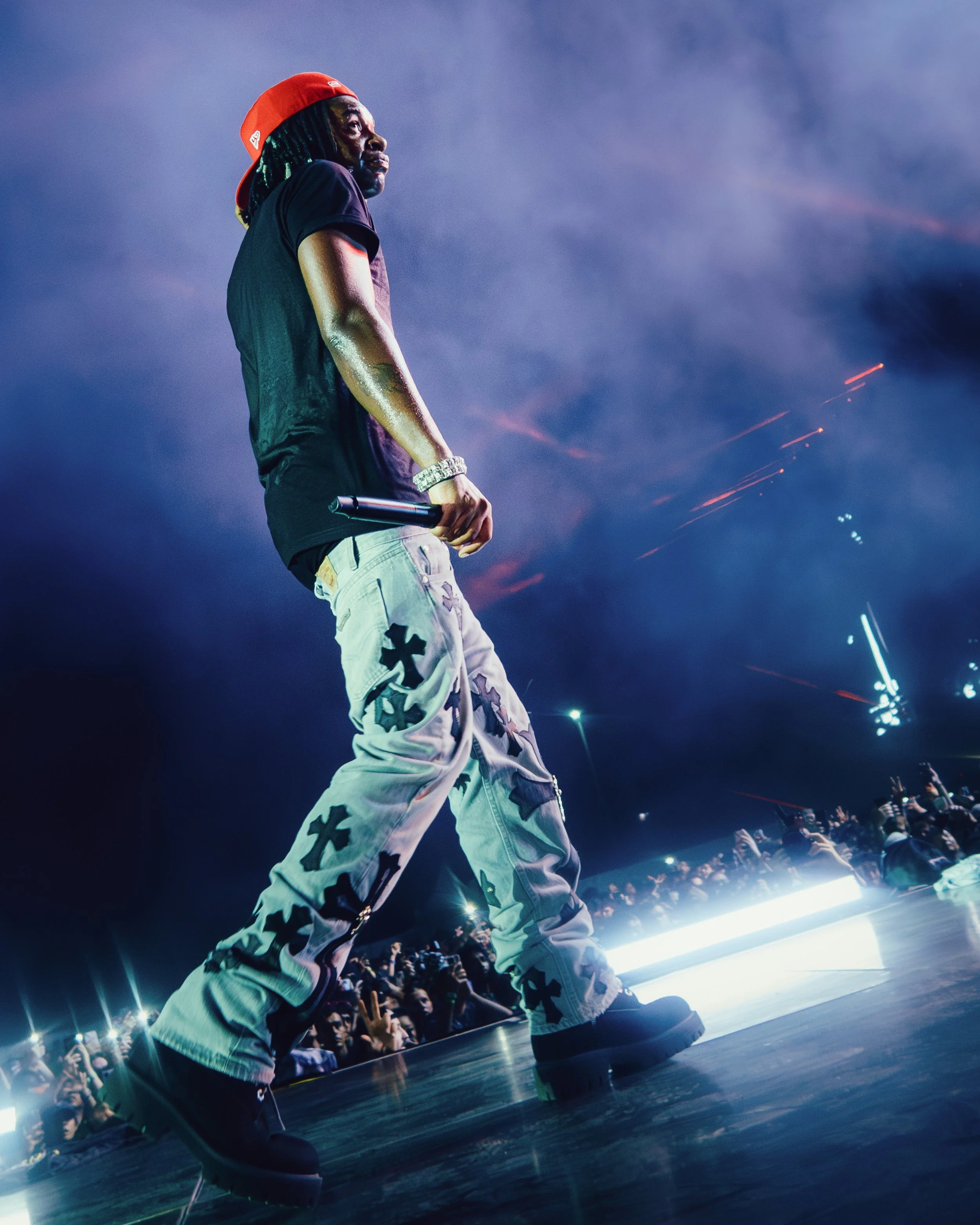 A male performer on stage during a concert, holding a microphone, wearing a red cap backwards and white pants with black cross patterns, with a crowd underneath and fireworks or pyrotechnics in the background.