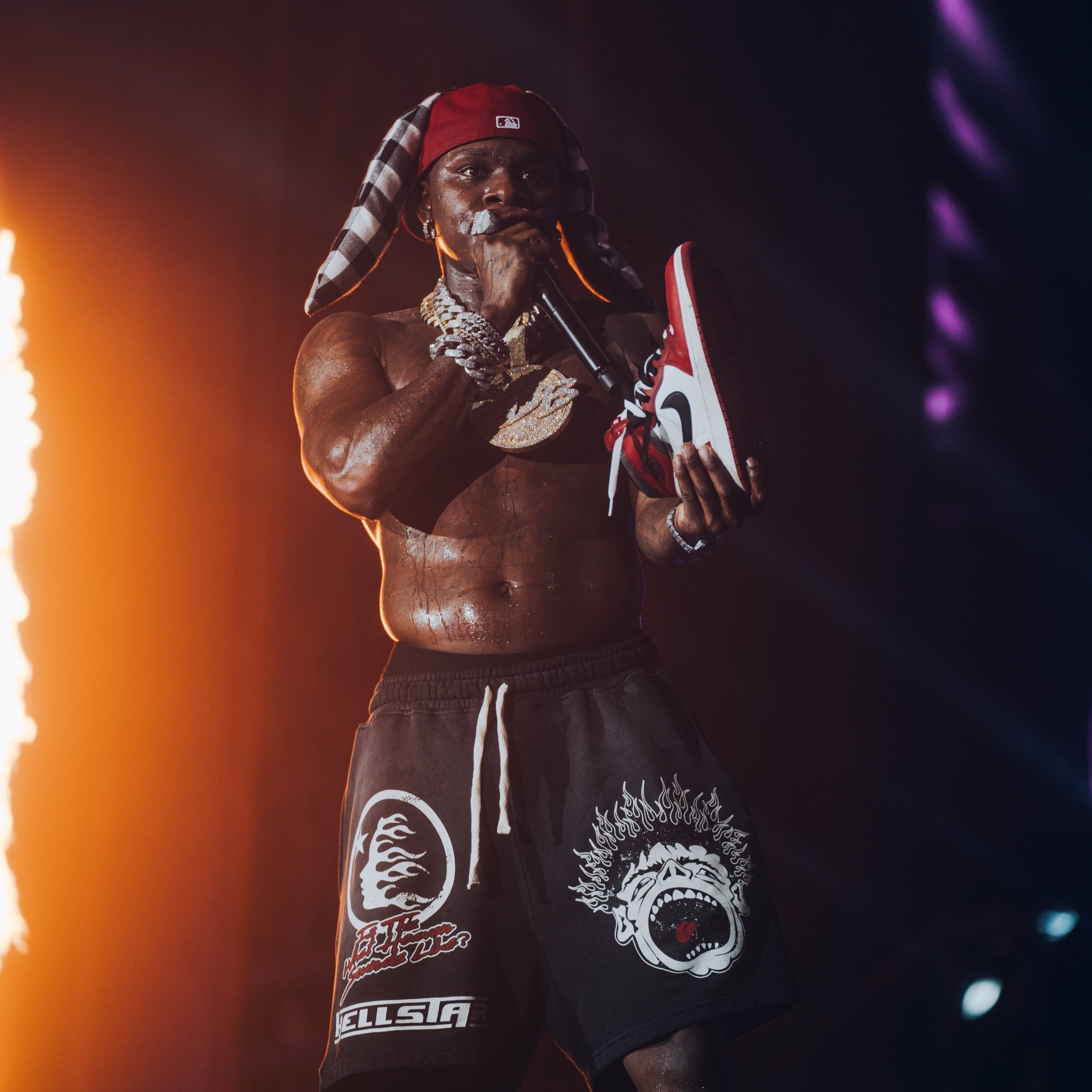 A shirtless man wearing a red cap backwards and a checkered scarf holds a sneaker and a microphone on stage with purple and orange lighting.