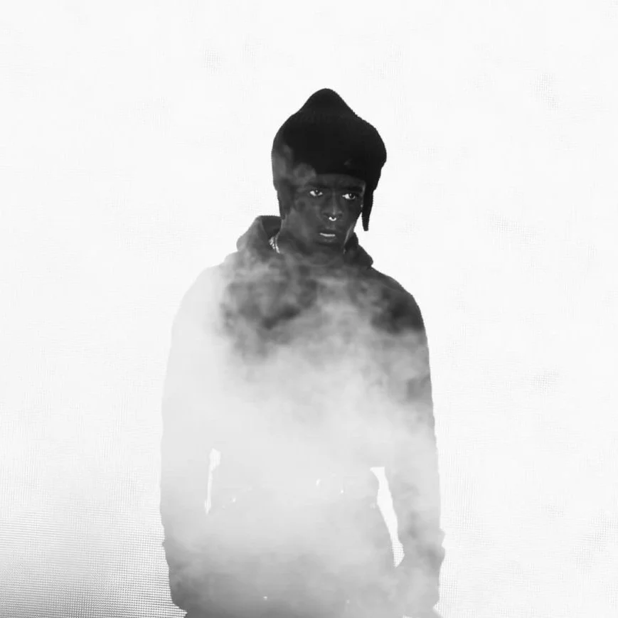 Black and white photo of a young person with dark skin, wearing a beanie and a nose ring, standing in front of a plain background with fog or smoke around their waist.