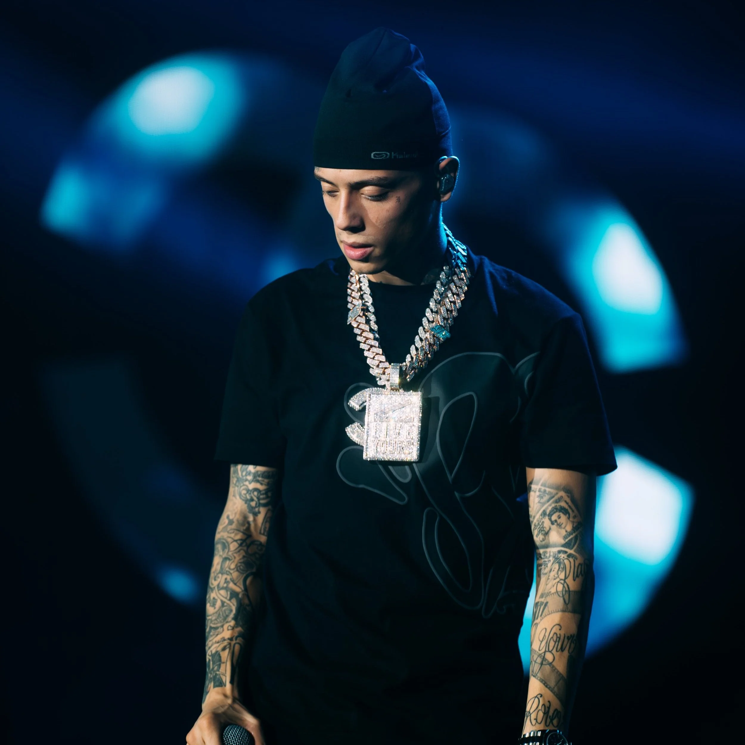 A young man wearing a black beanie, black t-shirt, and multiple large diamond-encrusted chains, standing on a stage with a dark background illuminated by blue lights.