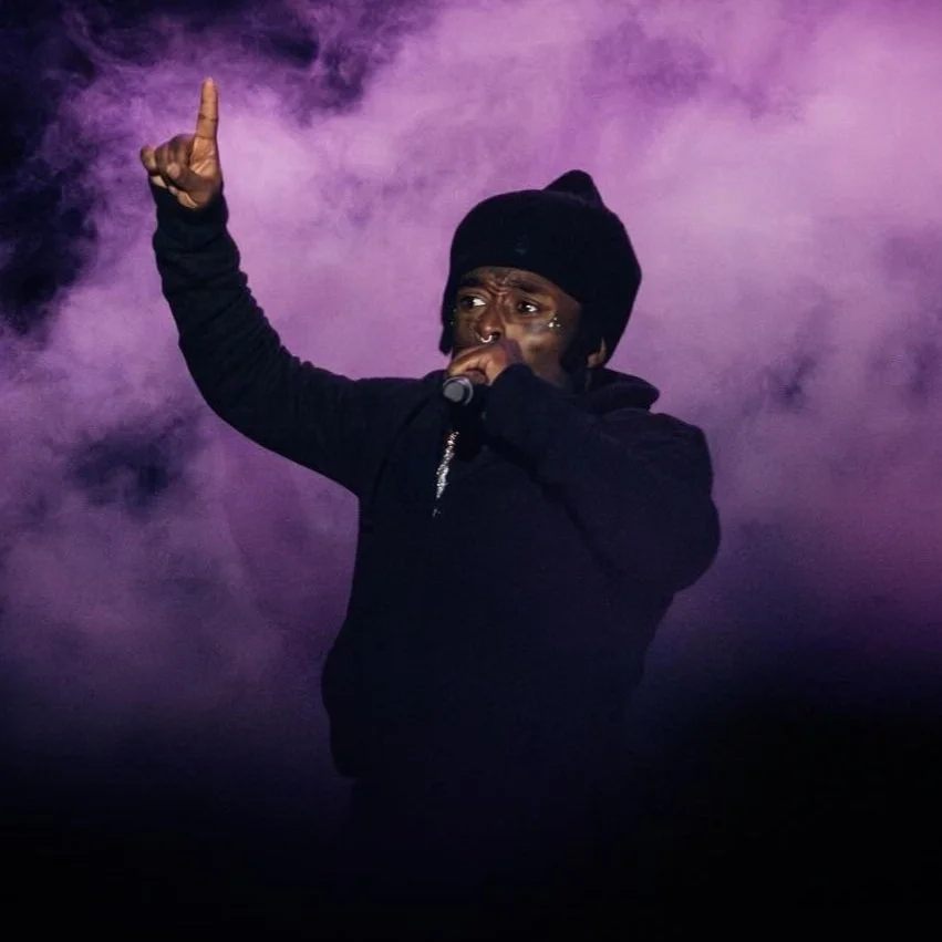 A performer on stage wearing a black beanie and black hoodie, holding a microphone, with purple stage smoke in the background.