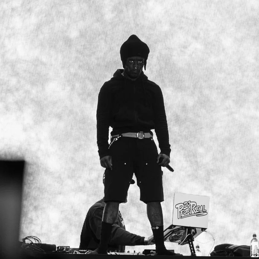 A man with dark skin dressed in a black hoodie and shorts, wearing a black beanie, standing on stage with a DJ and equipment in the background.