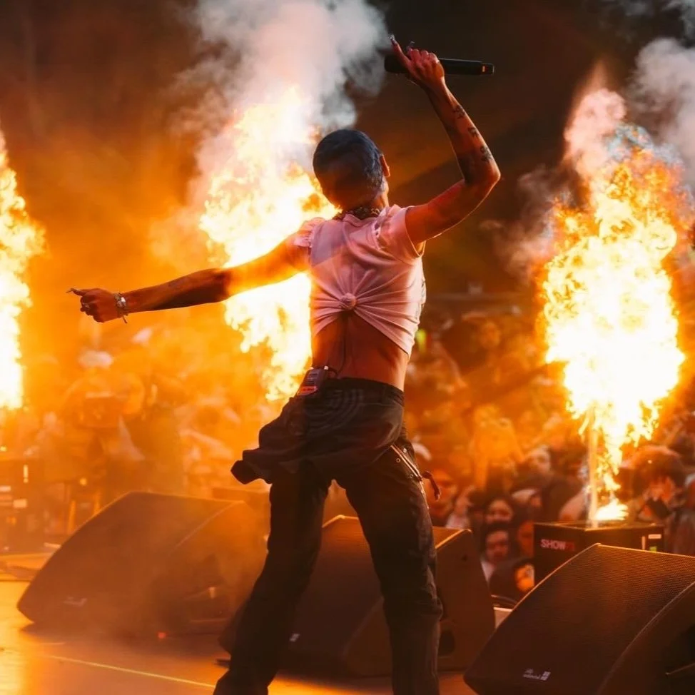A performer on stage holding a microphone with flames erupting in the background during a concert.