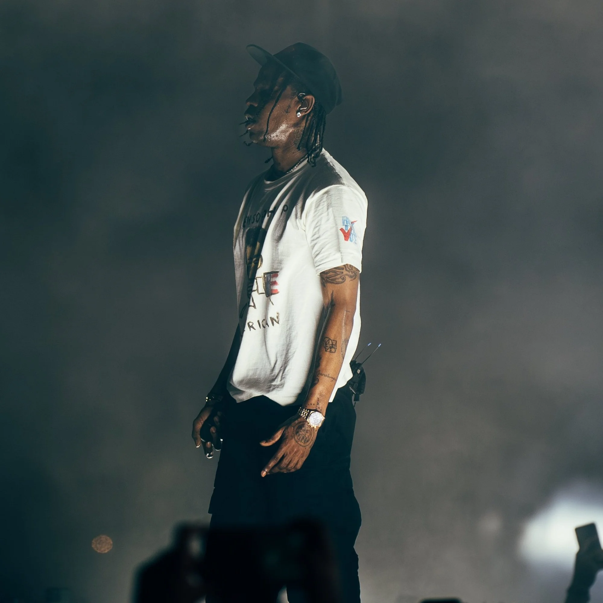 A man with tattoos on his arms wearing a white t-shirt, black pants, a cap, and jewelry, standing in a dark, smoky environment, holding an object in his left hand.