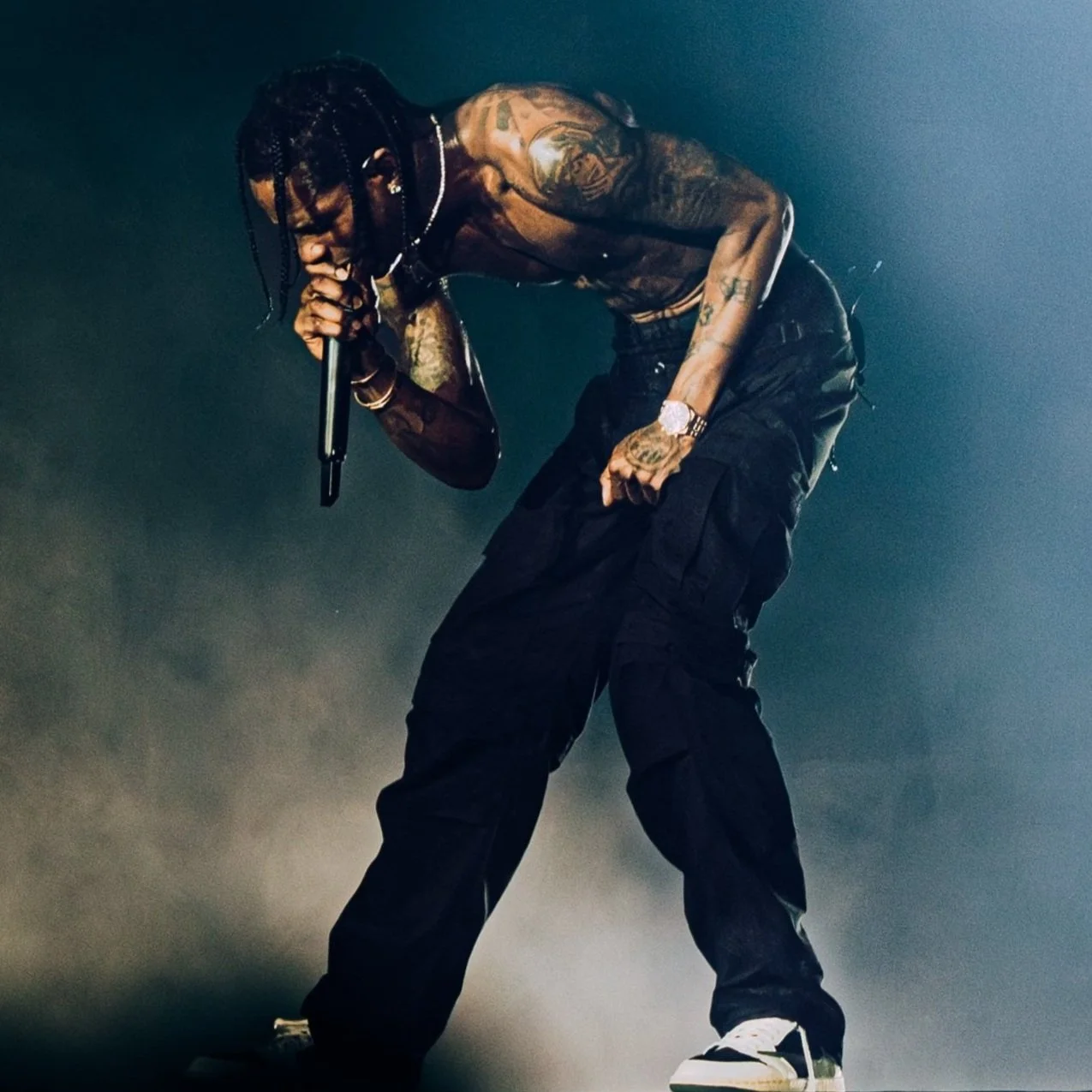 A male rapper performing on stage, holding a microphone close to his mouth, with tattoos on his arms, wearing black cargo pants, a watch, and white sneakers.
