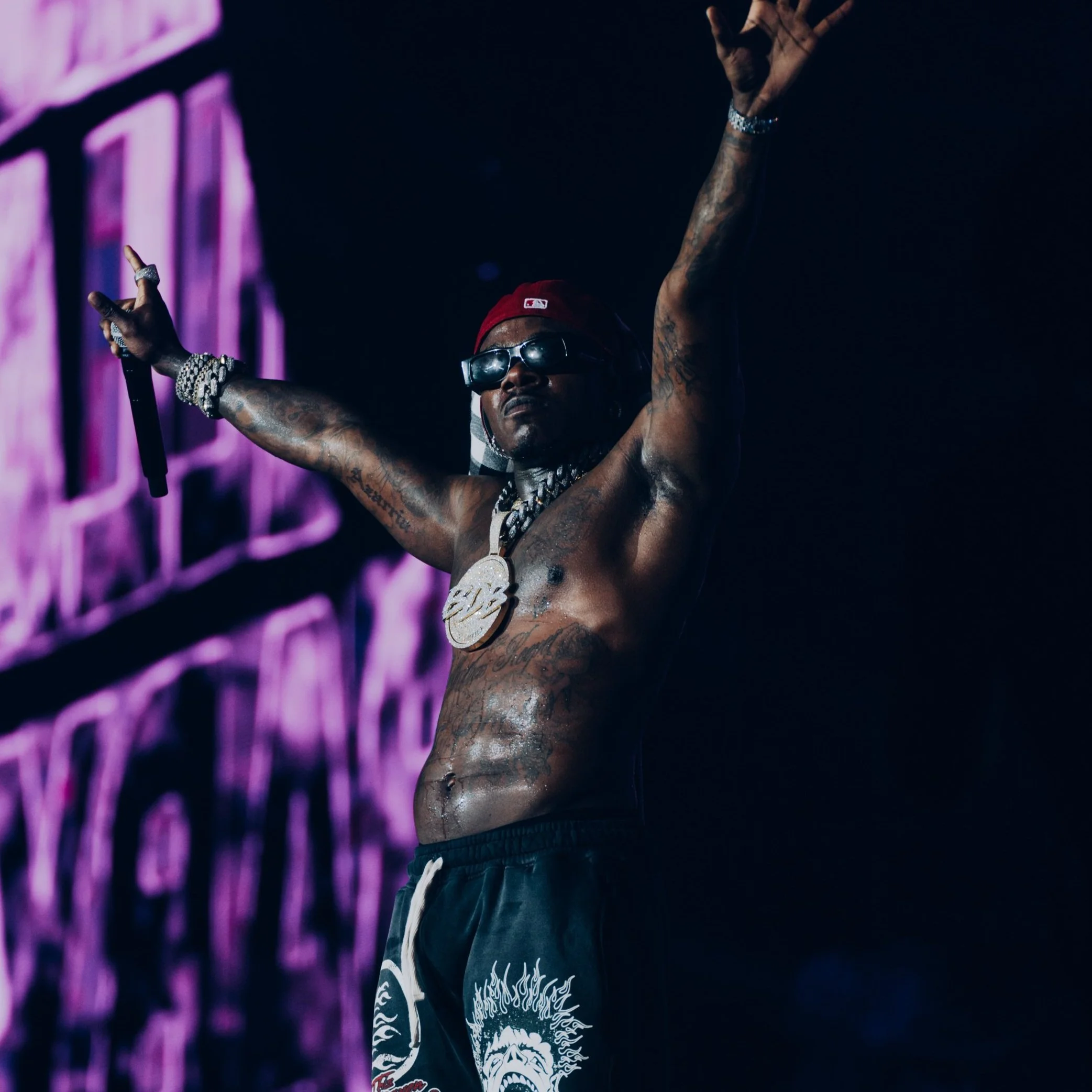 A shirtless man with tattoos, wearing dark sunglasses, a red cap, and jewelry, is on stage with his arms raised. There is a large screen behind him with purple visuals.