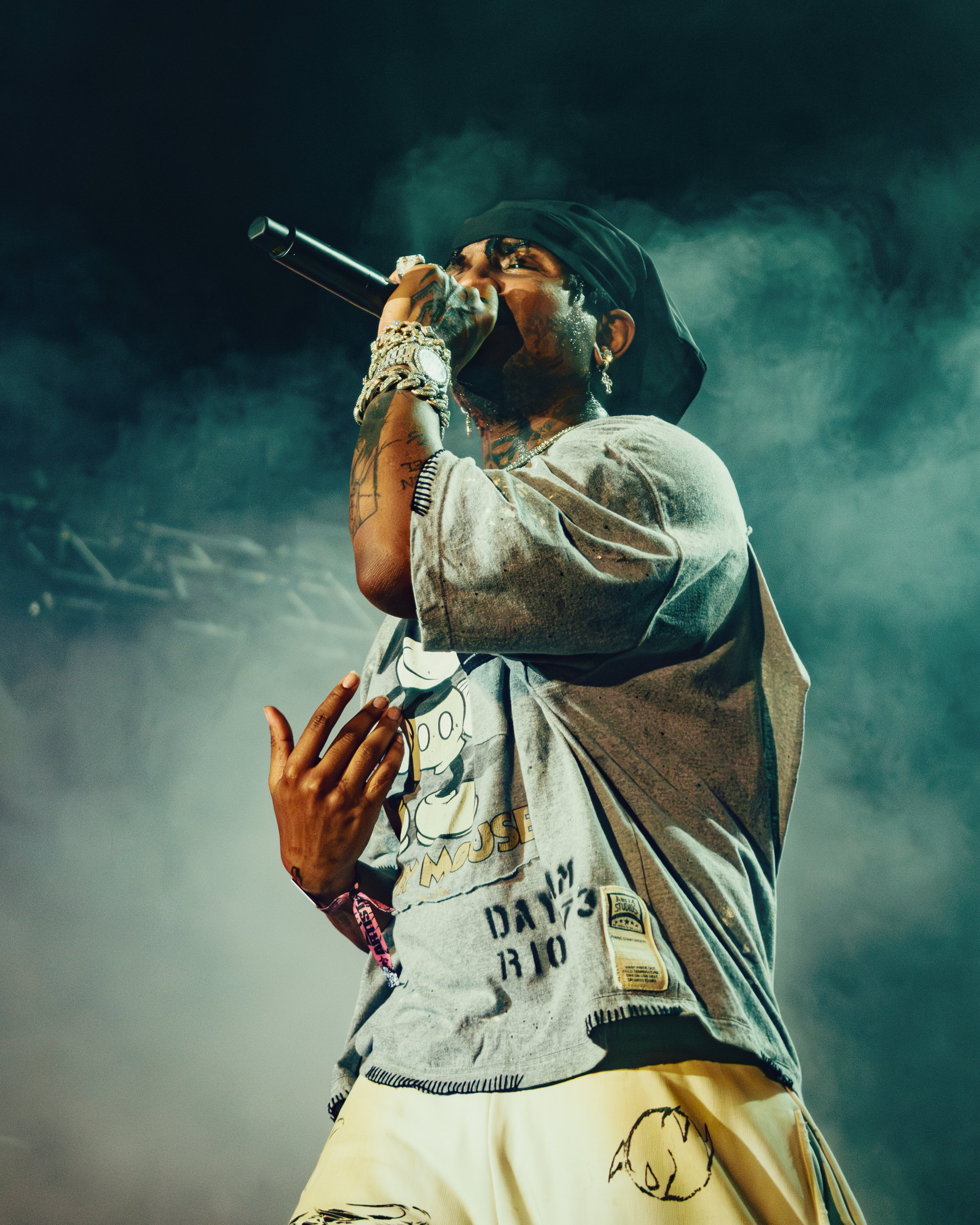 A male rapper performs on stage, holding a microphone close to his mouth with his hand adorned with jewelry, wearing a black headscarf, a graphic T-shirt, and light-colored pants.