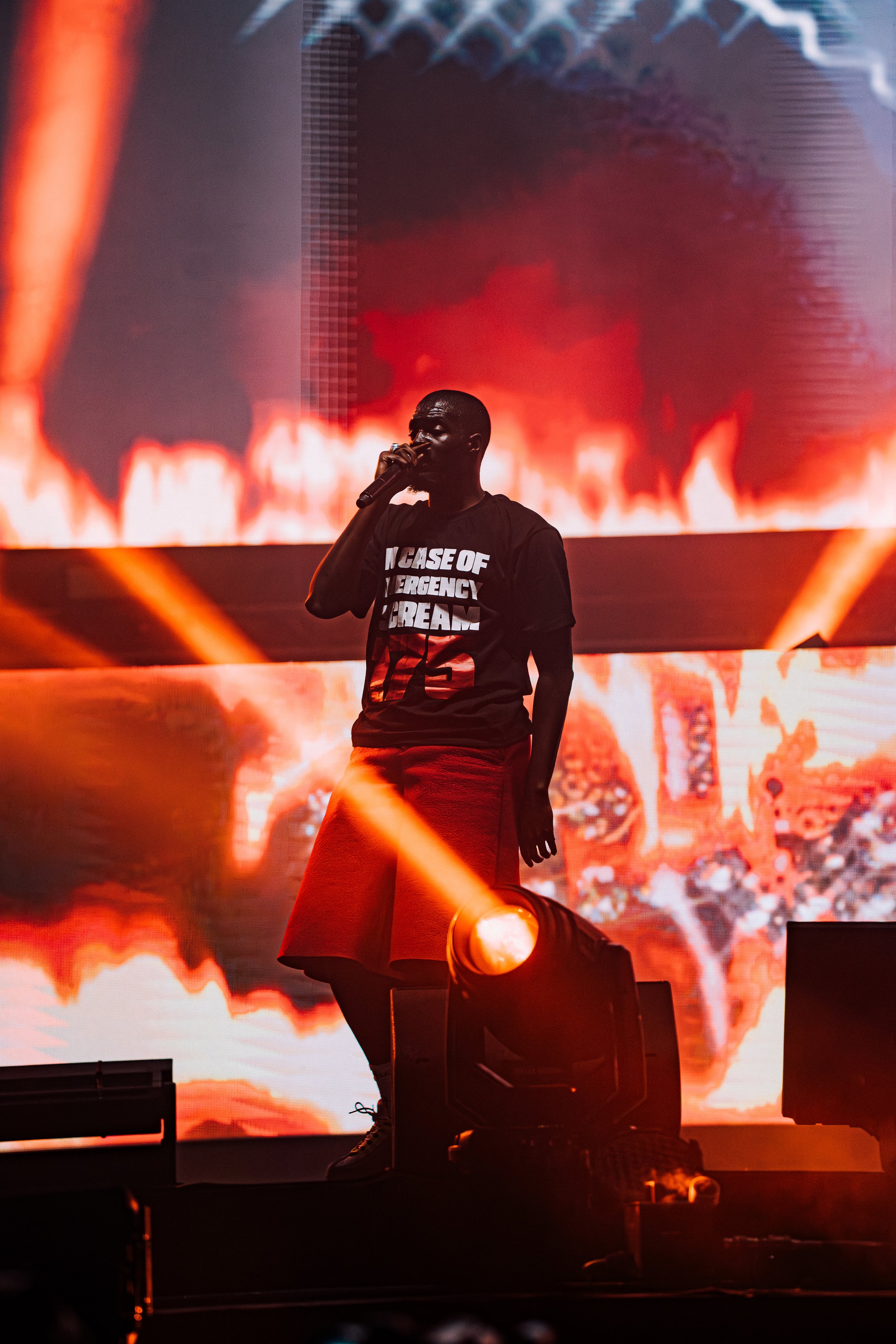 A performer stands on stage holding a microphone with bright red and orange lighting and digital flames on a large screen behind them.