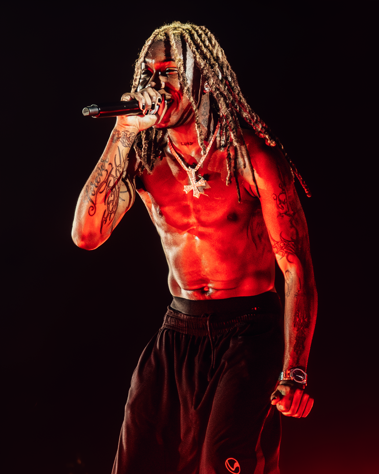 A shirtless male performer with dreadlocks holding a microphone on stage, illuminated by red and yellow lighting.