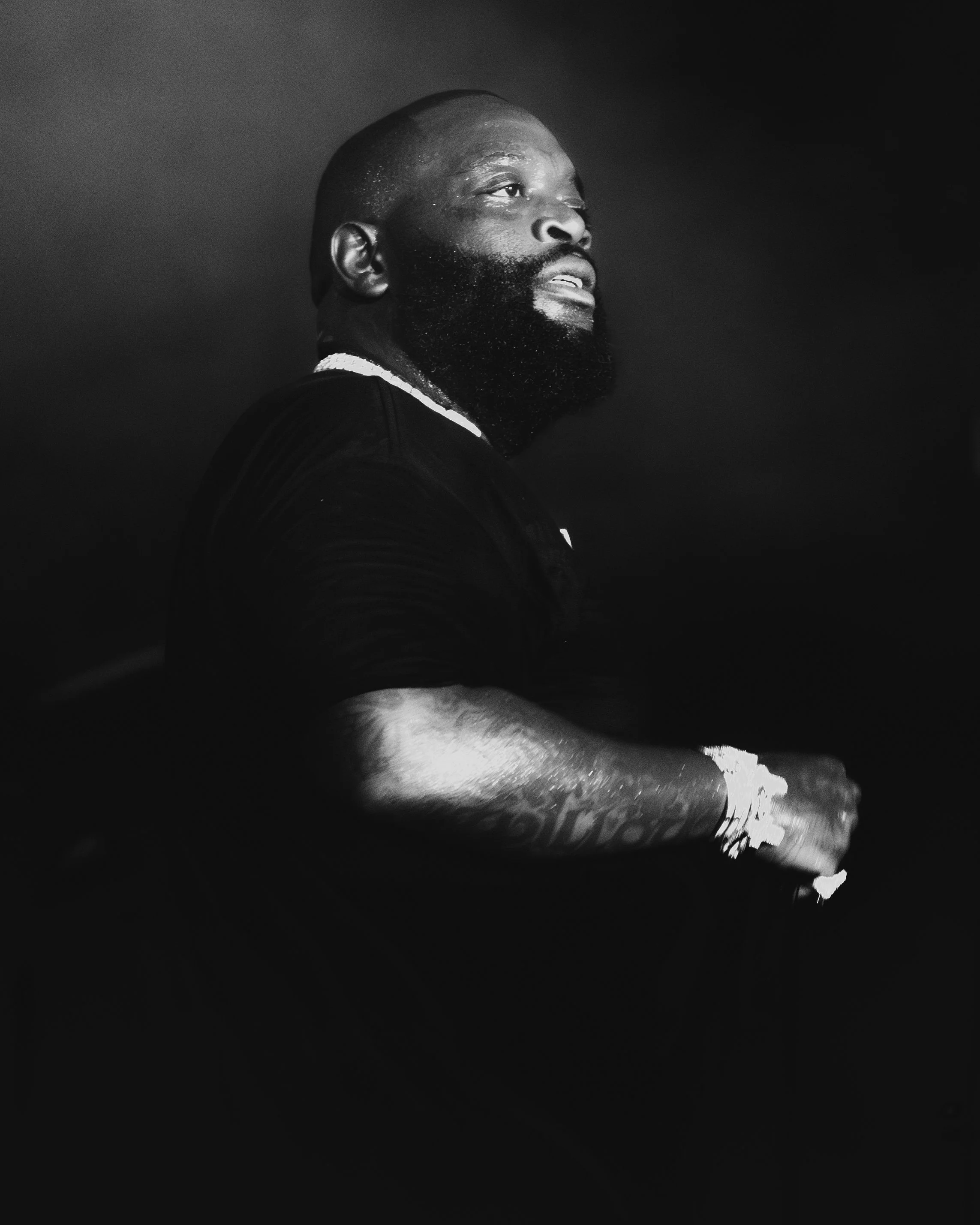 Black and white portrait of a bearded man with a shaved head, looking to the side, dressed in black, with water droplets on his arm and face.