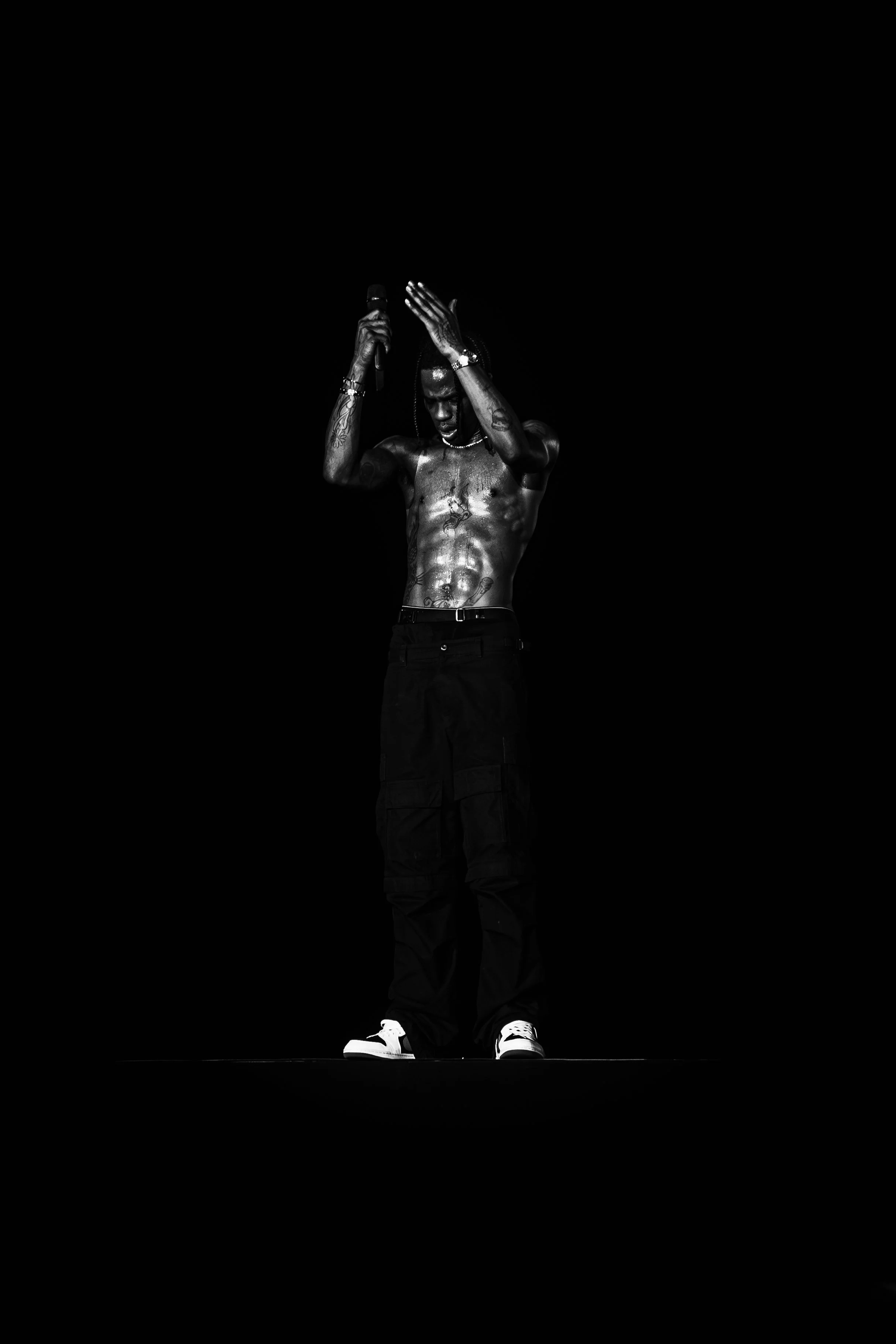 A shirtless man with tattoos, wearing black pants and white sneakers, stands against a black background, holding a microphone in one hand and touching his head with the other.