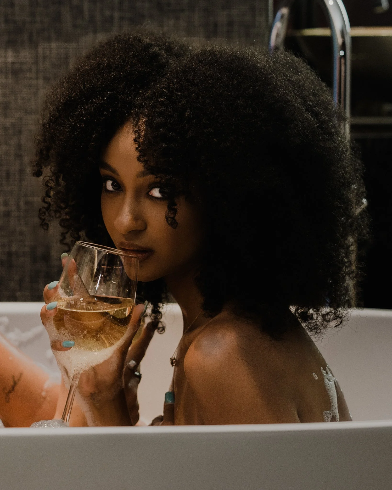 A woman with curly hair taking a bath and holding a glass of sparkling wine, looking at the camera.